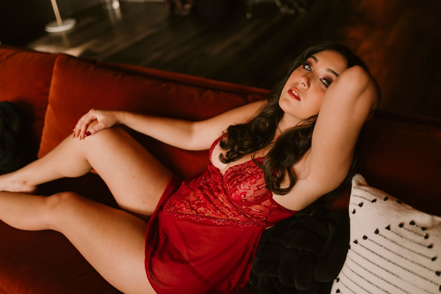Woman in red lace dress reclining on a velvet sofa in a moody, dimly lit room.