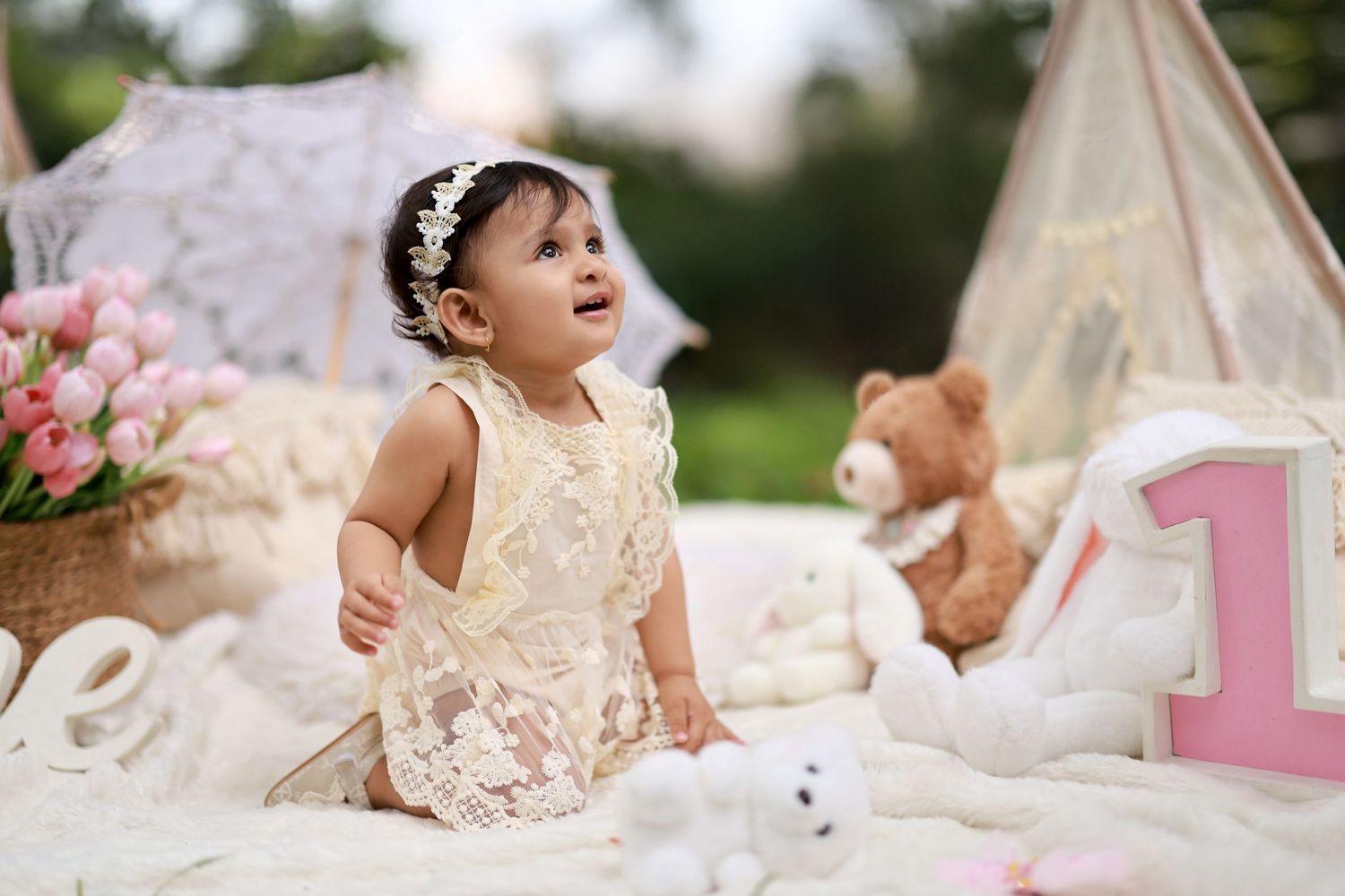 A baby girl in a lace dress sits outdoors at a first birthday setup with a teddy bear and pink number one.