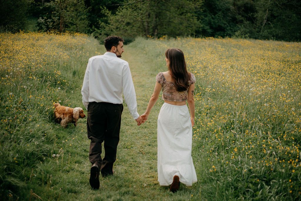 A couple walks hand in hand through a wildflower meadow with their golden cocker spaniel.