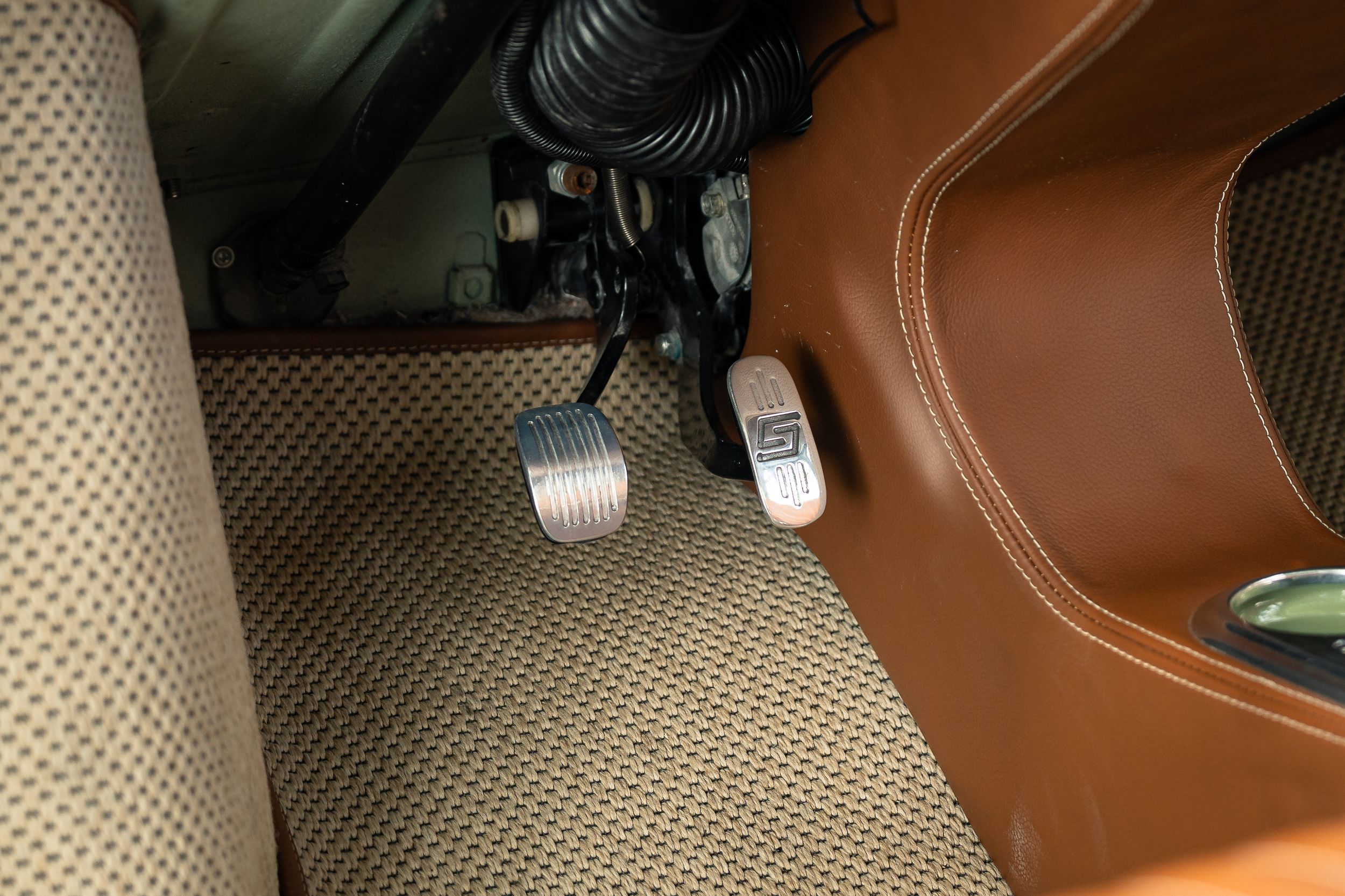 Close-up of brake and accelerator pedals on tan leather floor mat in a restored classic car interior.