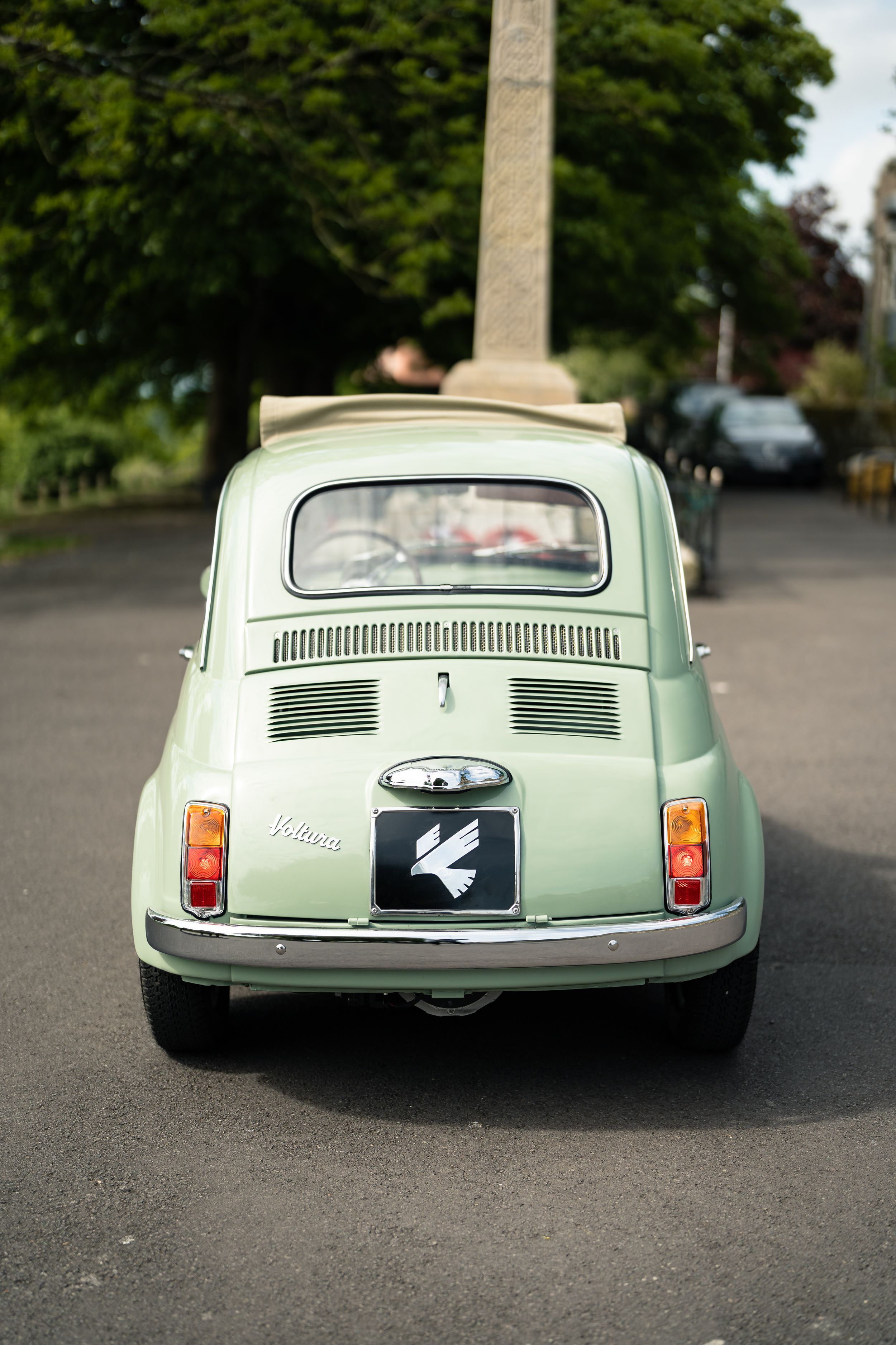Rear view of a mint green Fiat 500 Nuova with Viottaz badge driving on a tree-lined road.