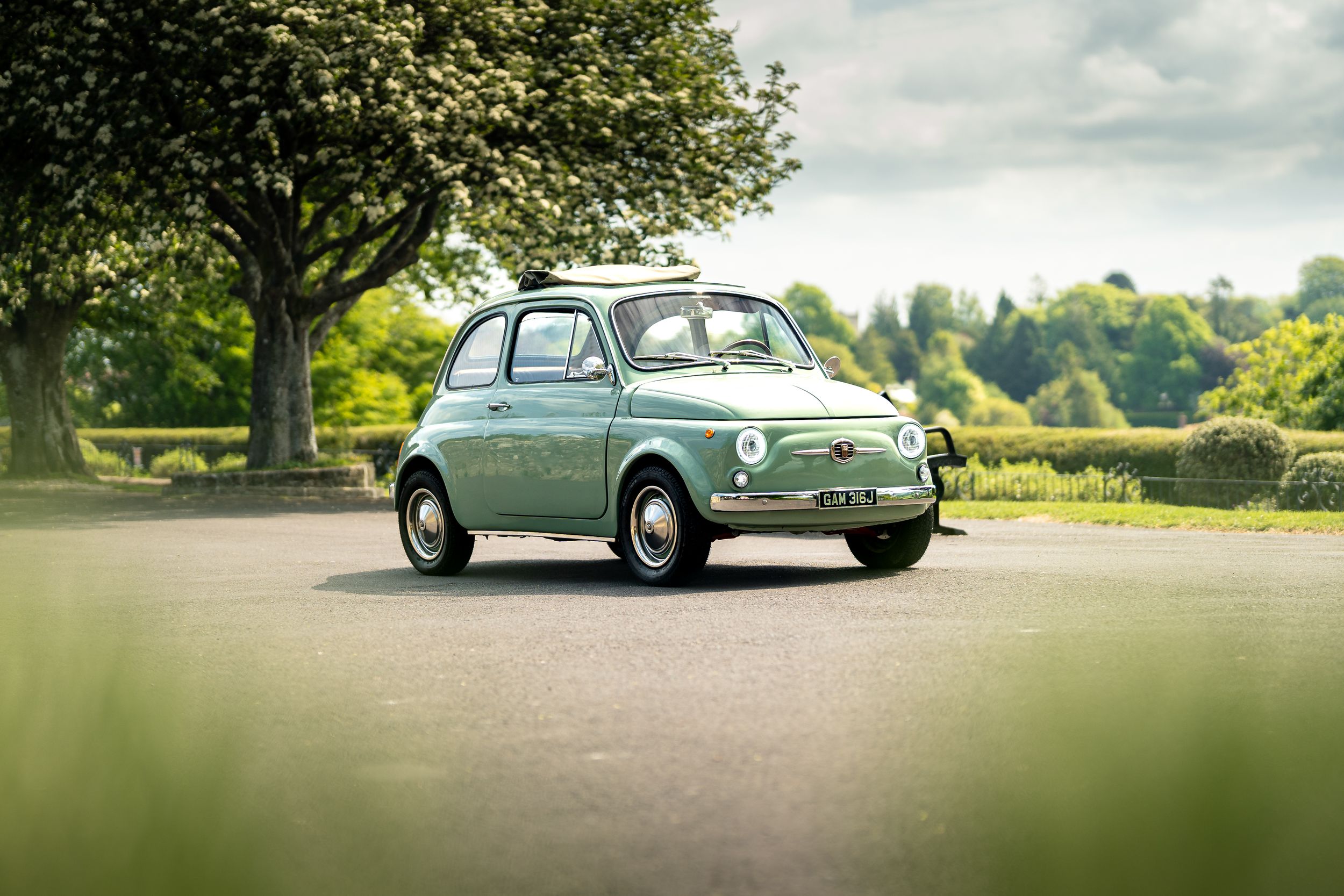 Restored mint green classic Fiat 500 parked on a tree-lined driveway in lush green surroundings.