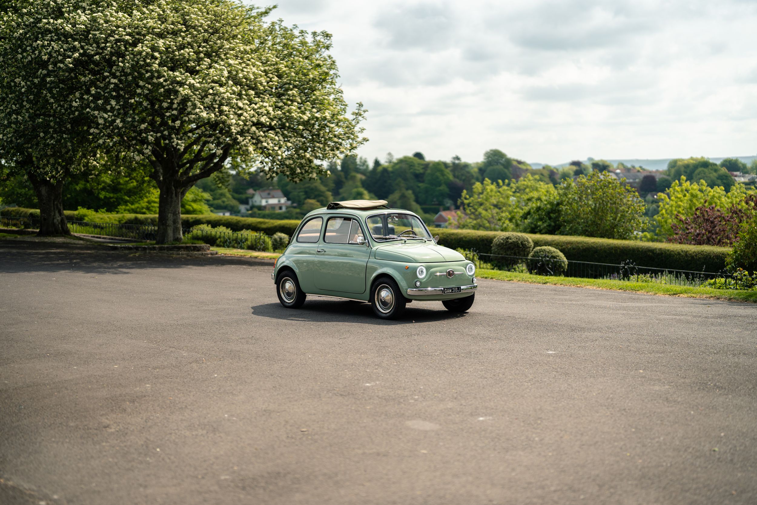 Vintage mint green Fiat 500 classic car parked on open tarmac surrounded by lush green trees.