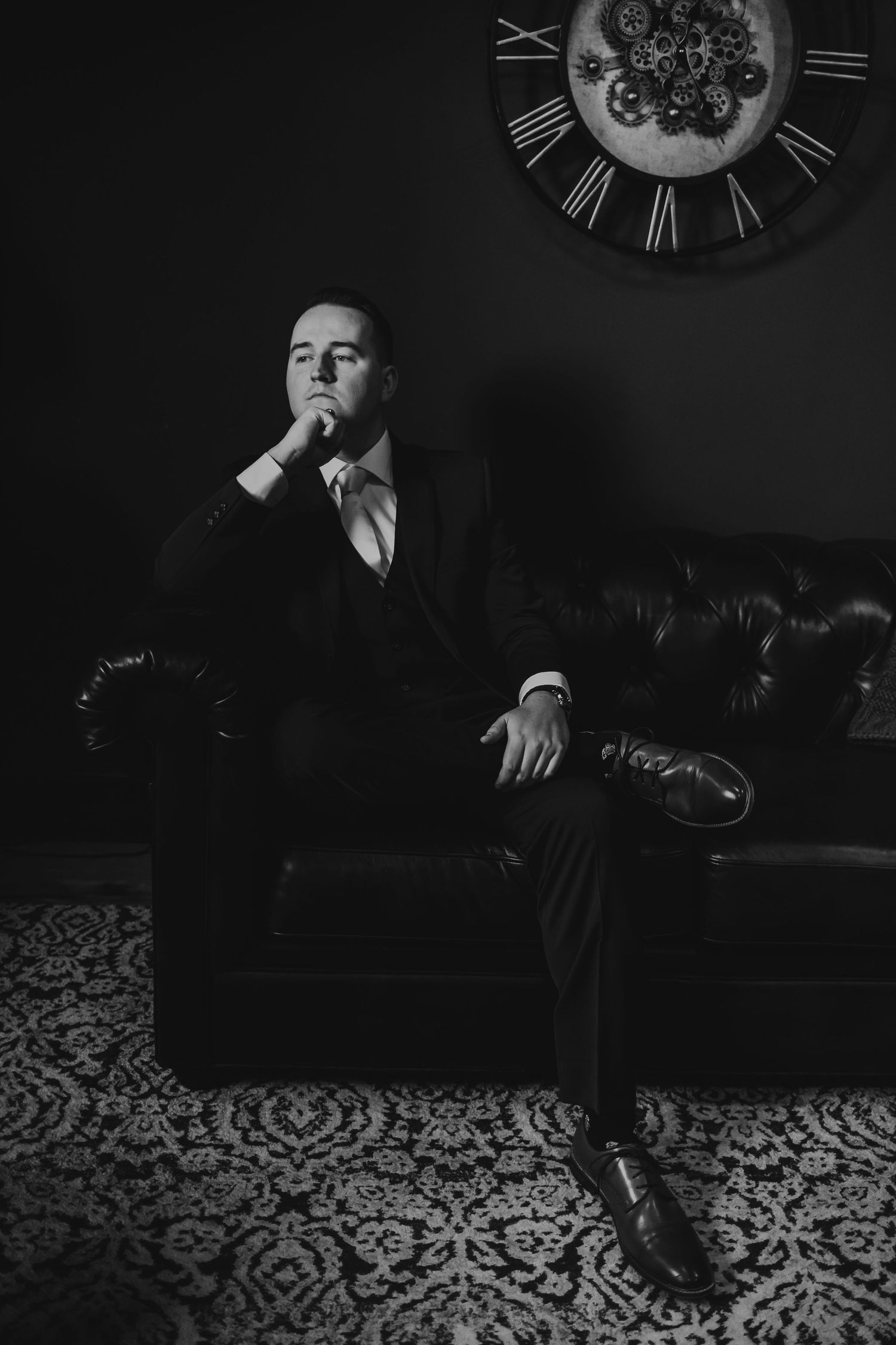 A man in a suit sits pensively on a leather sofa in a dark room with a decorative clock on the wall.