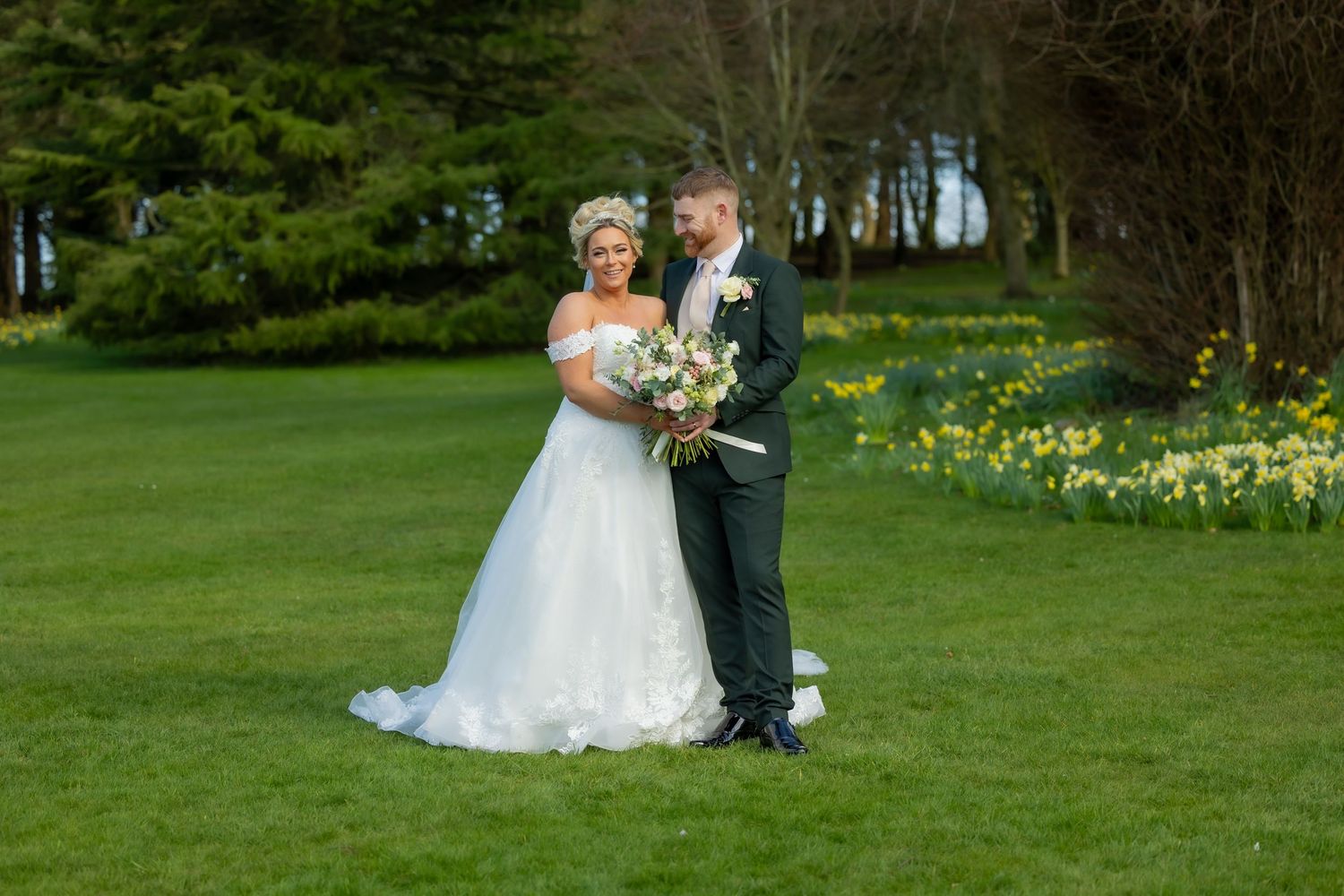 Bride and groom posing together on lush green lawn surrounded by daffodils and trees on their spring wedding day. Wedding Casual Captures