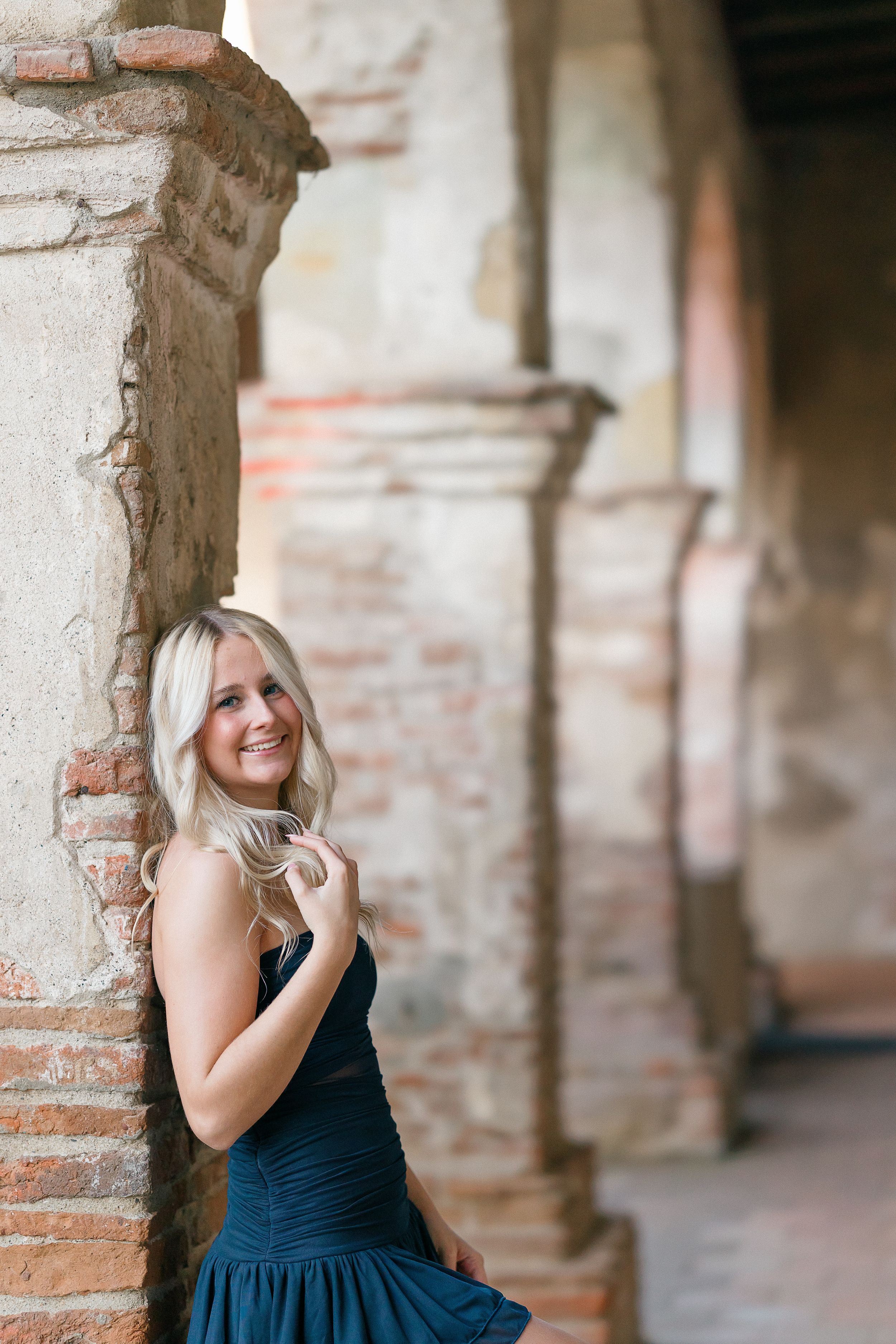 Woman in navy blue dress poses thoughtfully against rustic brick archways in an outdoor setting.
