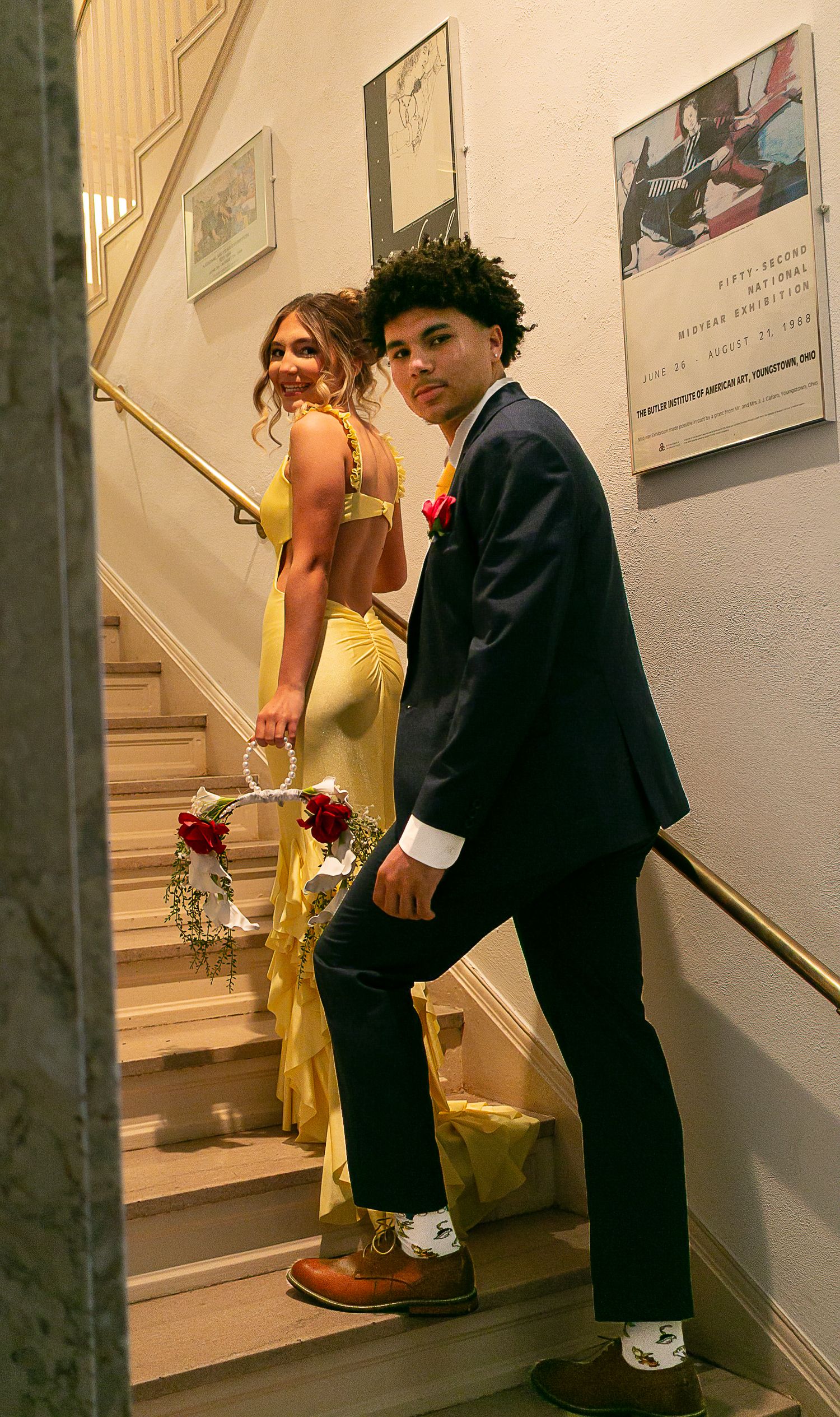 A young couple dressed in prom attire pose on a staircase, she in yellow gown, he in dark suit with boutonniere.