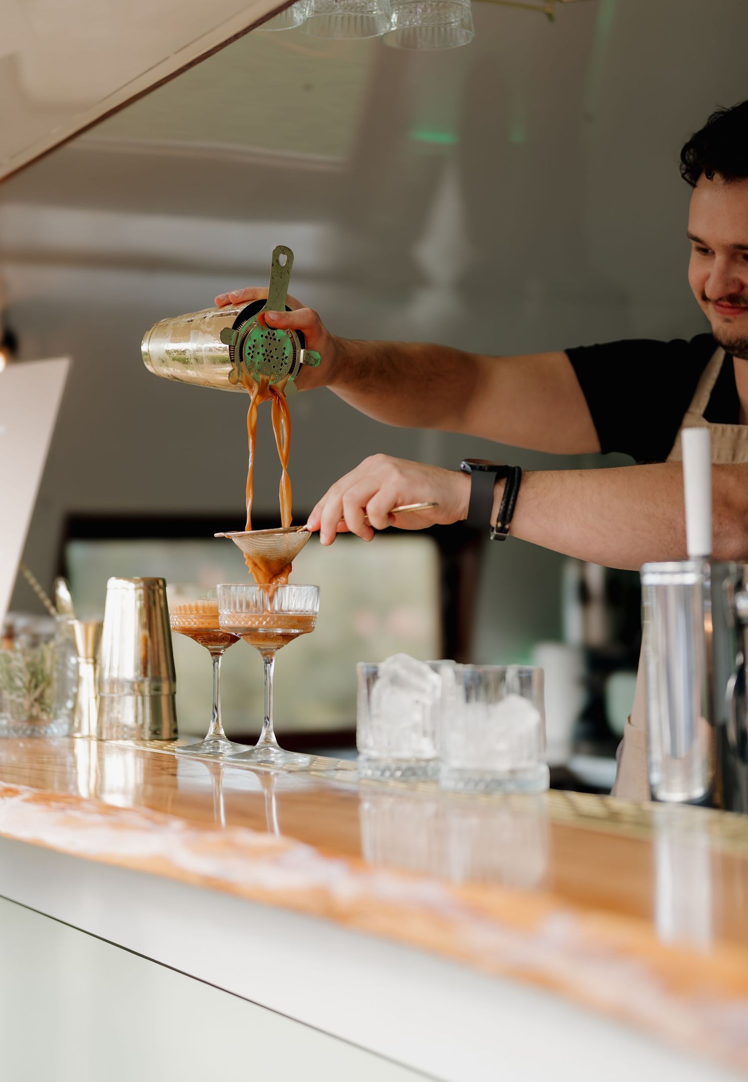 Bartender pouring espresso martini cocktails through a strainer at a stylish bar counter with ice and shakers.
