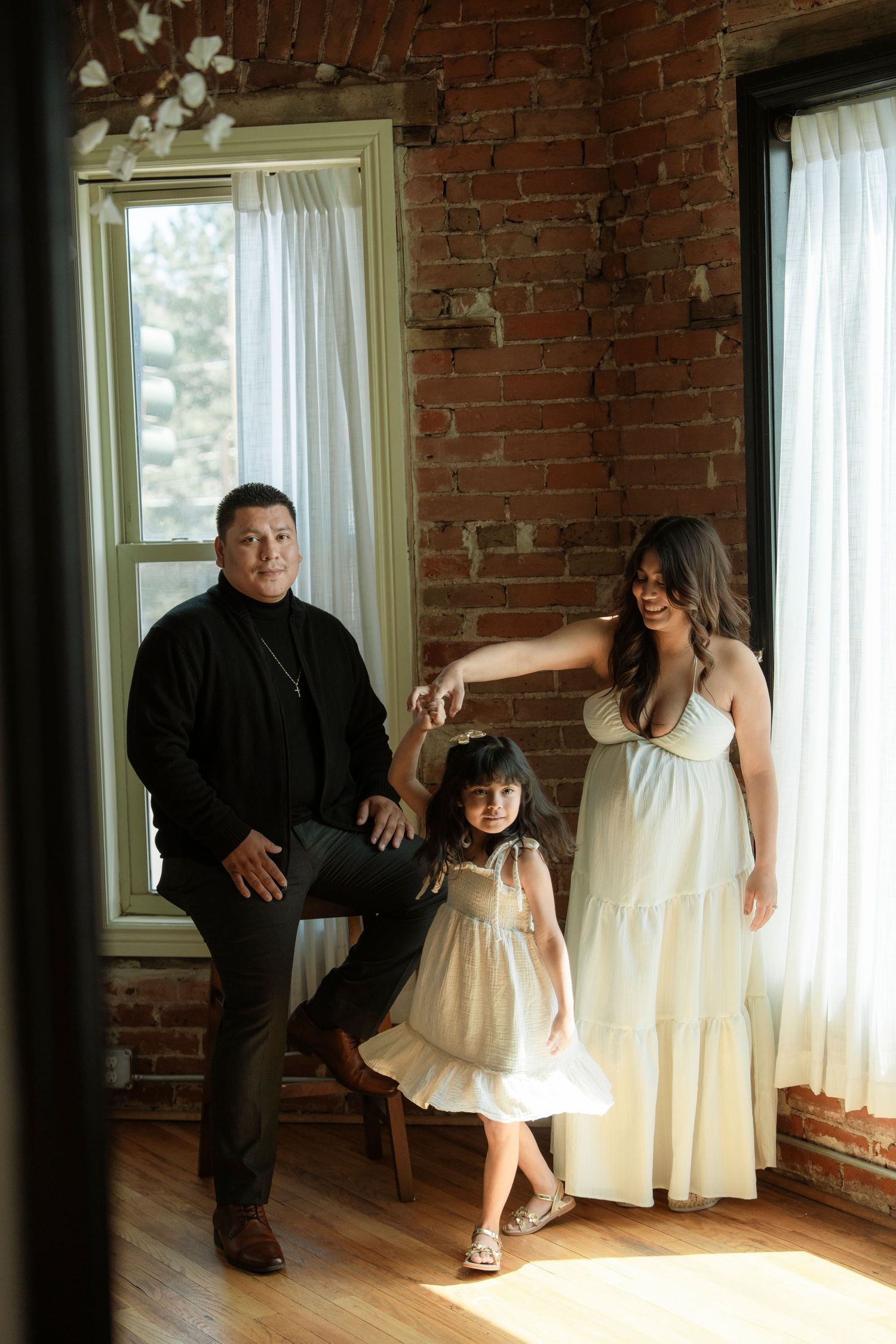 Family of three posing in sunlit brick-walled room, parents holding toddler girl's hands as she twirls in white dress.