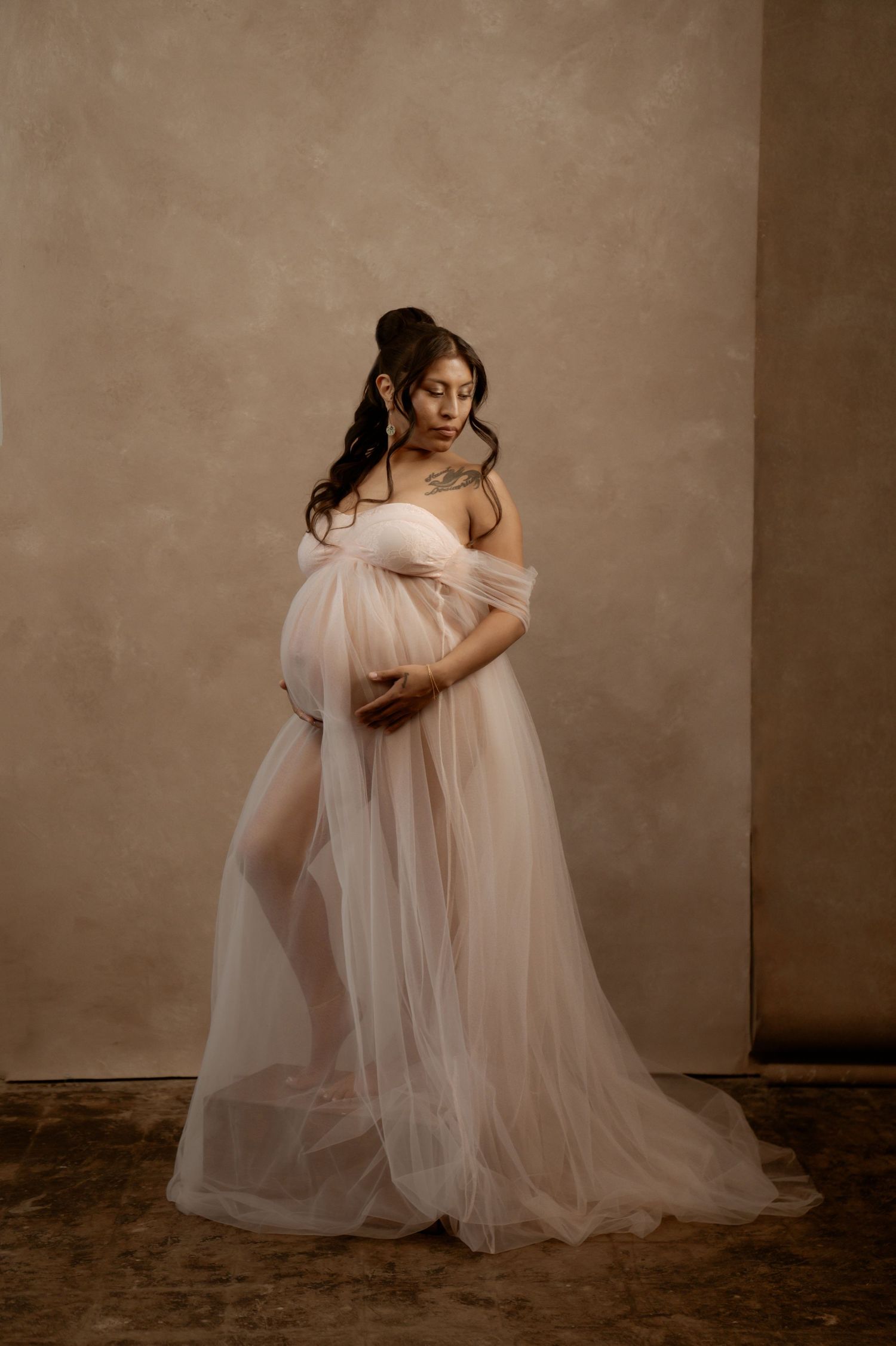 Pregnant woman in flowing blush tulle gown cradling baby bump in elegant studio maternity portrait.