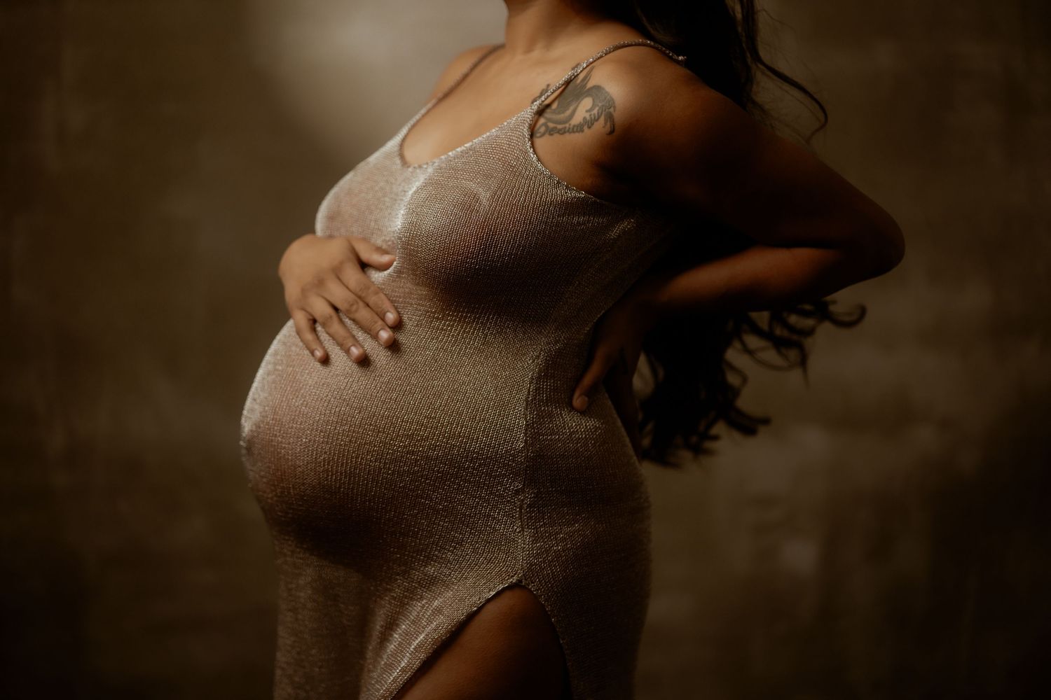 Pregnant woman in a shimmering beige dress with tattoos, cradling her baby bump in a moody studio setting.