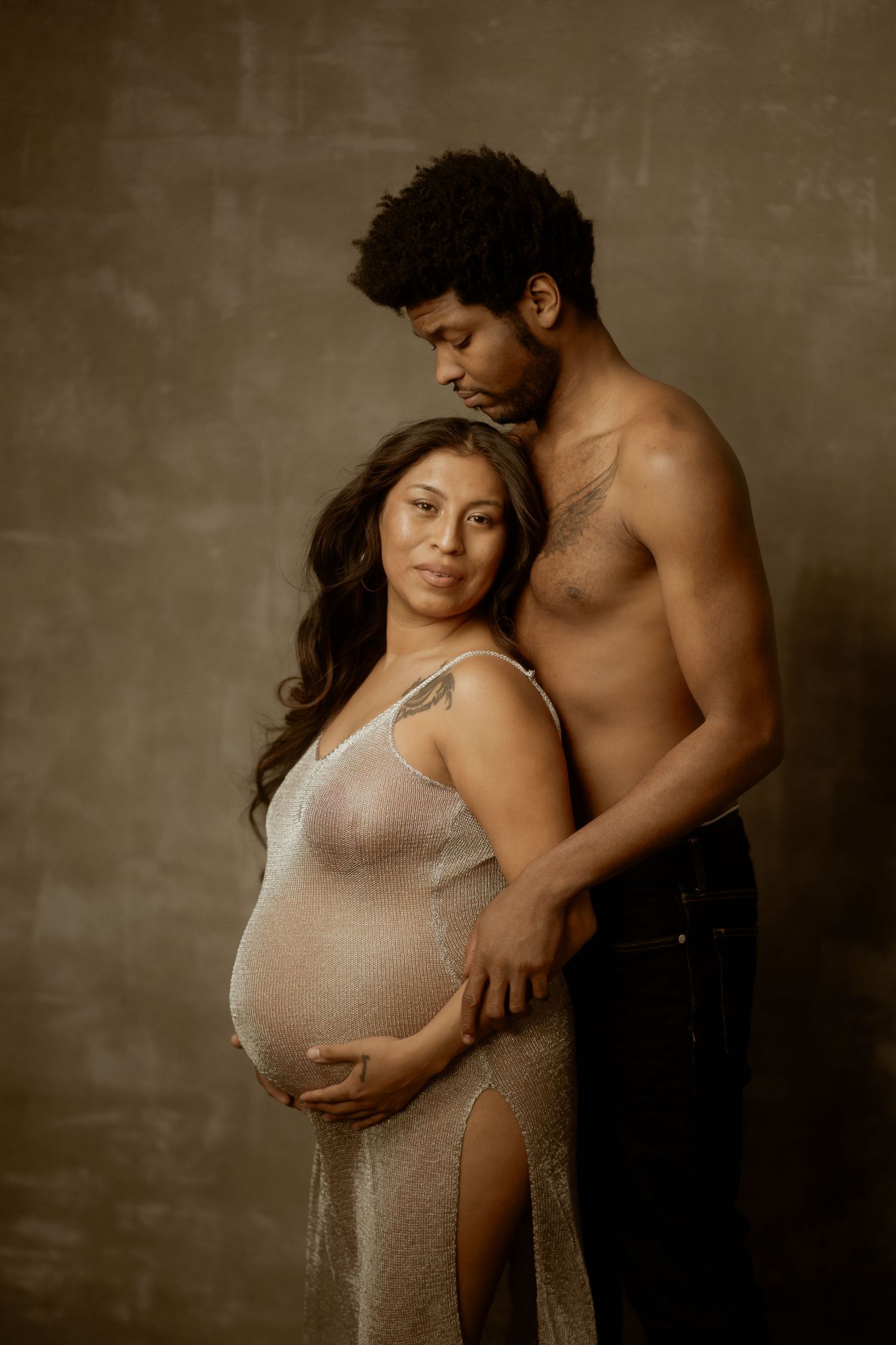 Pregnant woman in sheer dress cradling her baby bump while her partner embraces her in a studio maternity portrait.