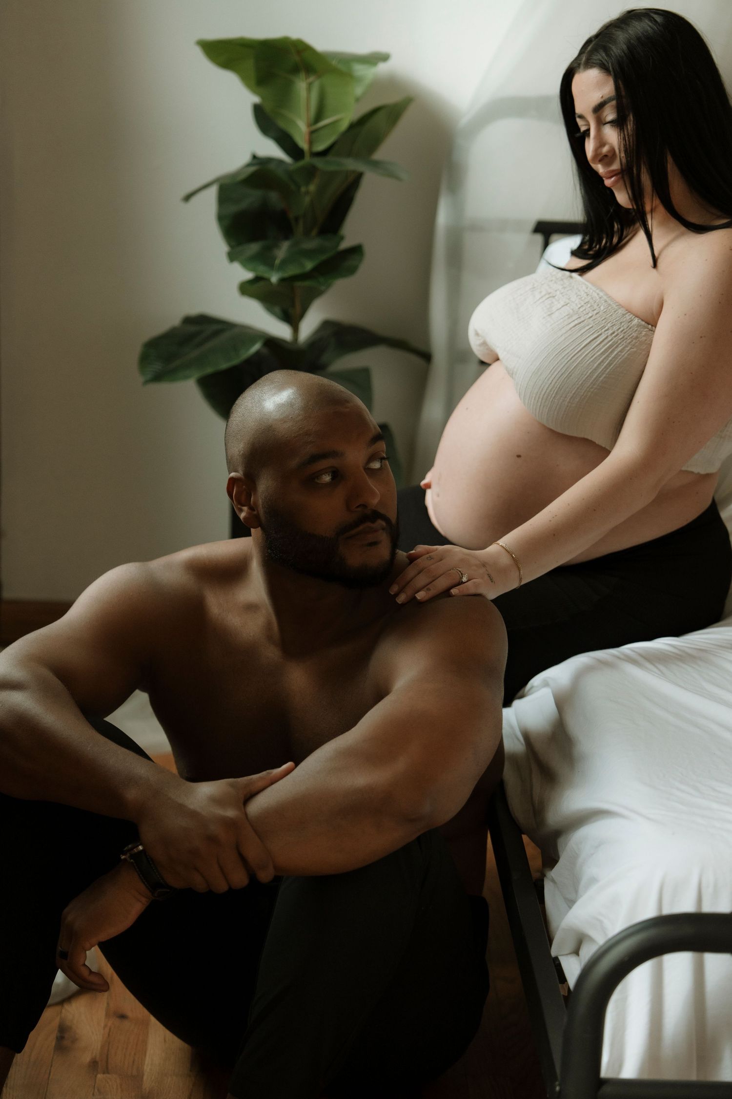 A shirtless man sits on the floor gazing up at a pregnant woman in a white crop top during an intimate maternity photo session.