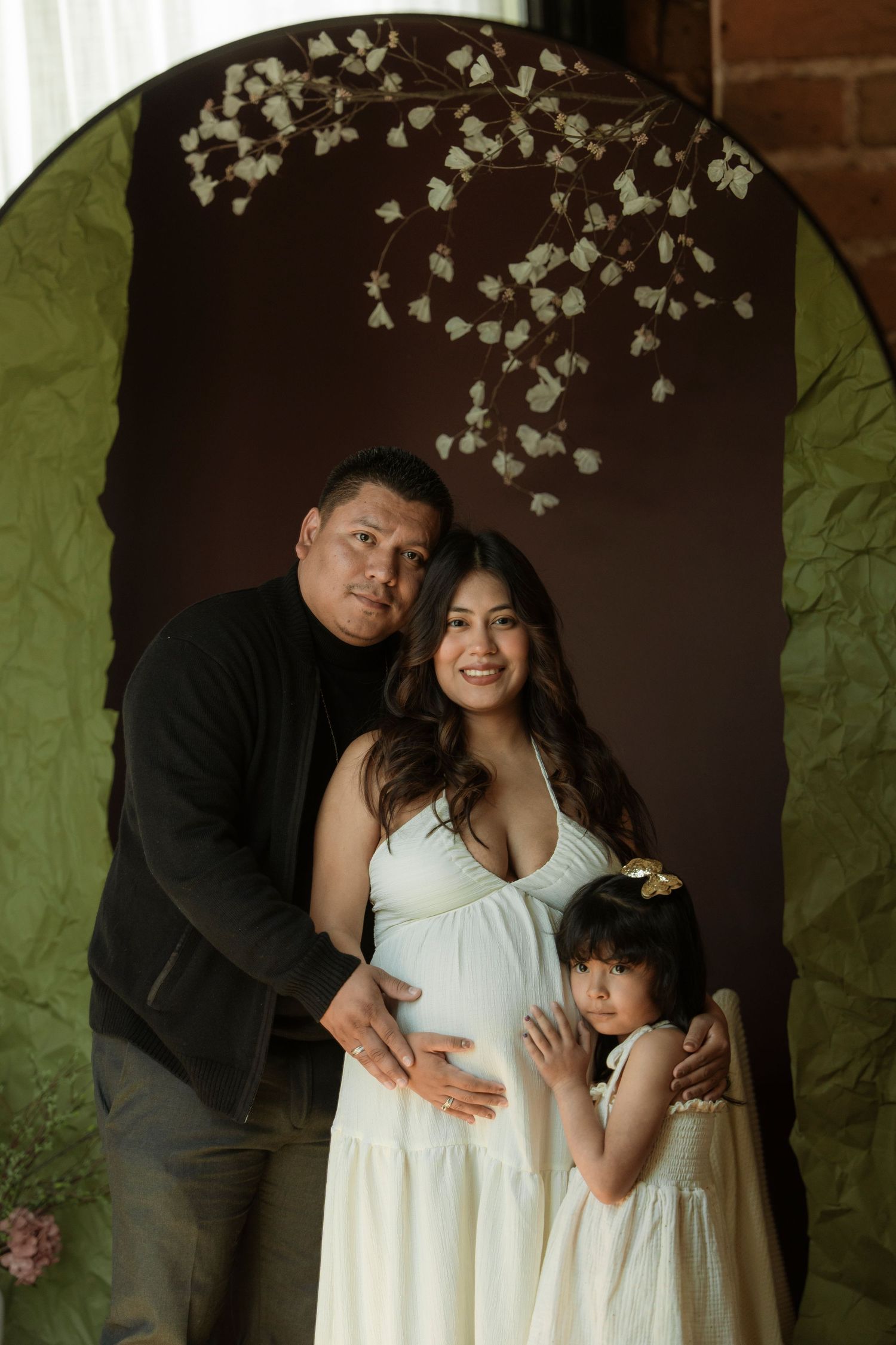 Family maternity photo with pregnant woman in white dress, man in black, and young girl posing under floral arch.