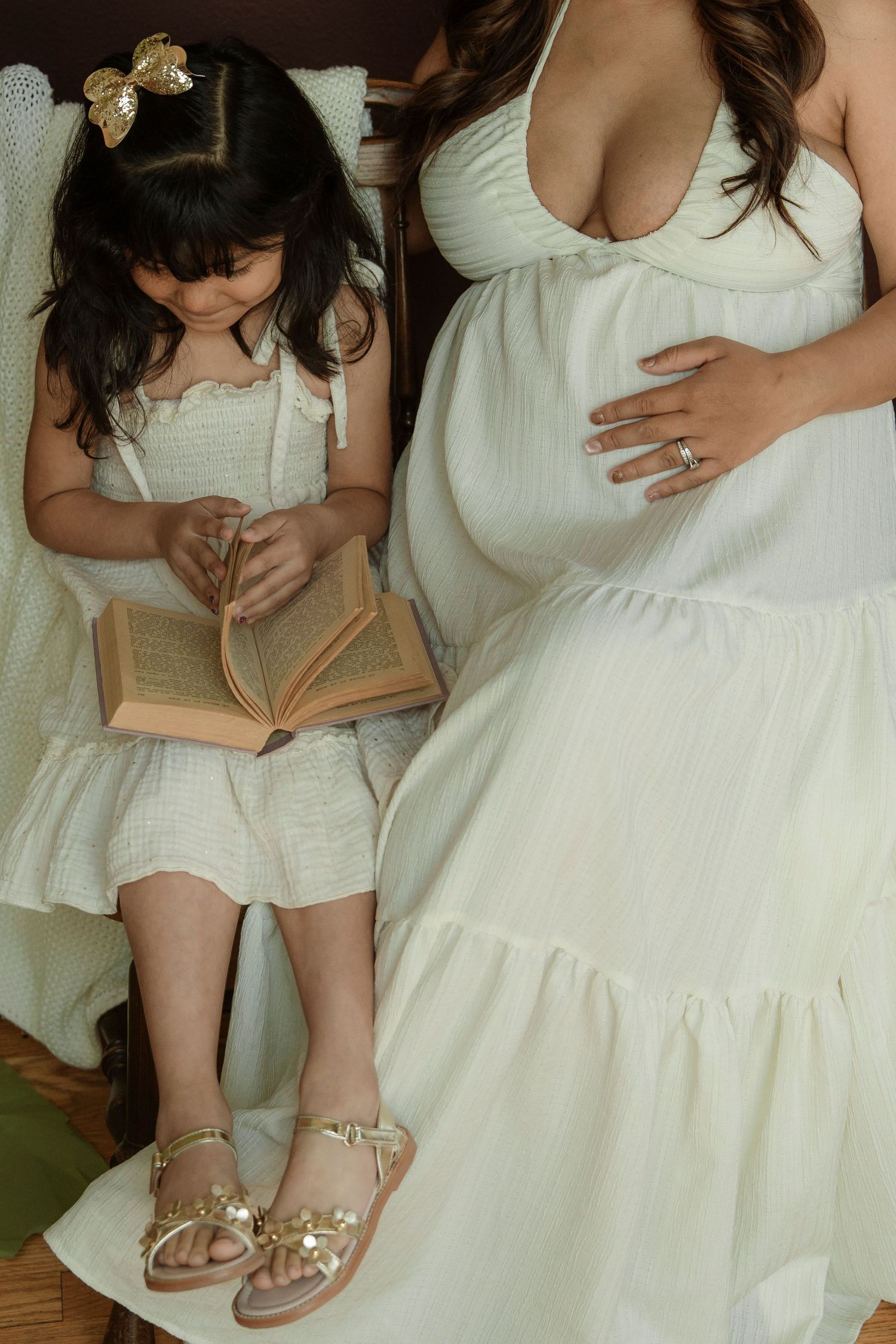 Pregnant woman in white dress sits beside young girl in matching outfit reading a book, both wearing gold accessories.
