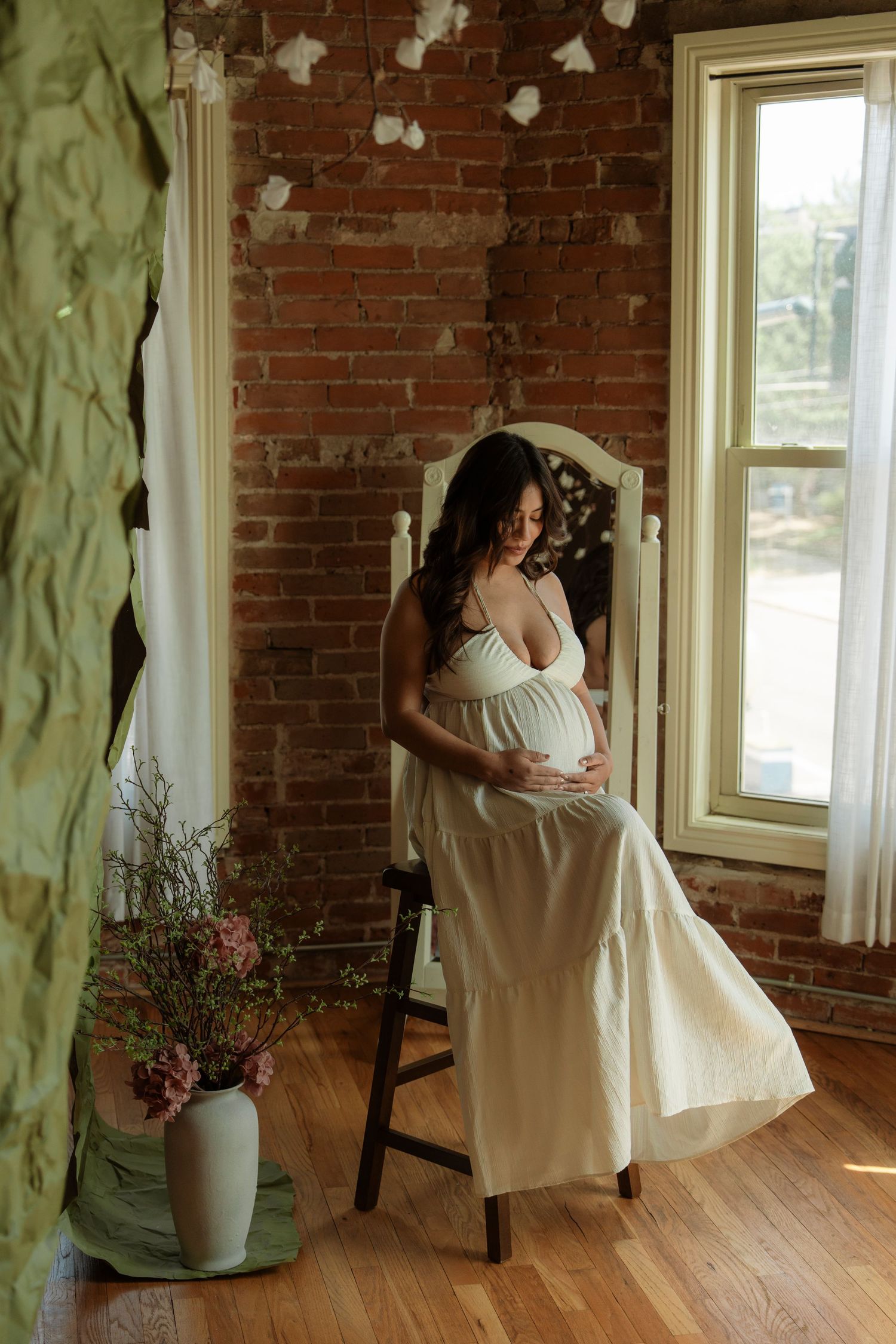 Pregnant woman in white dress cradling belly near mirror, brick wall, and window in a warmly lit studio.
