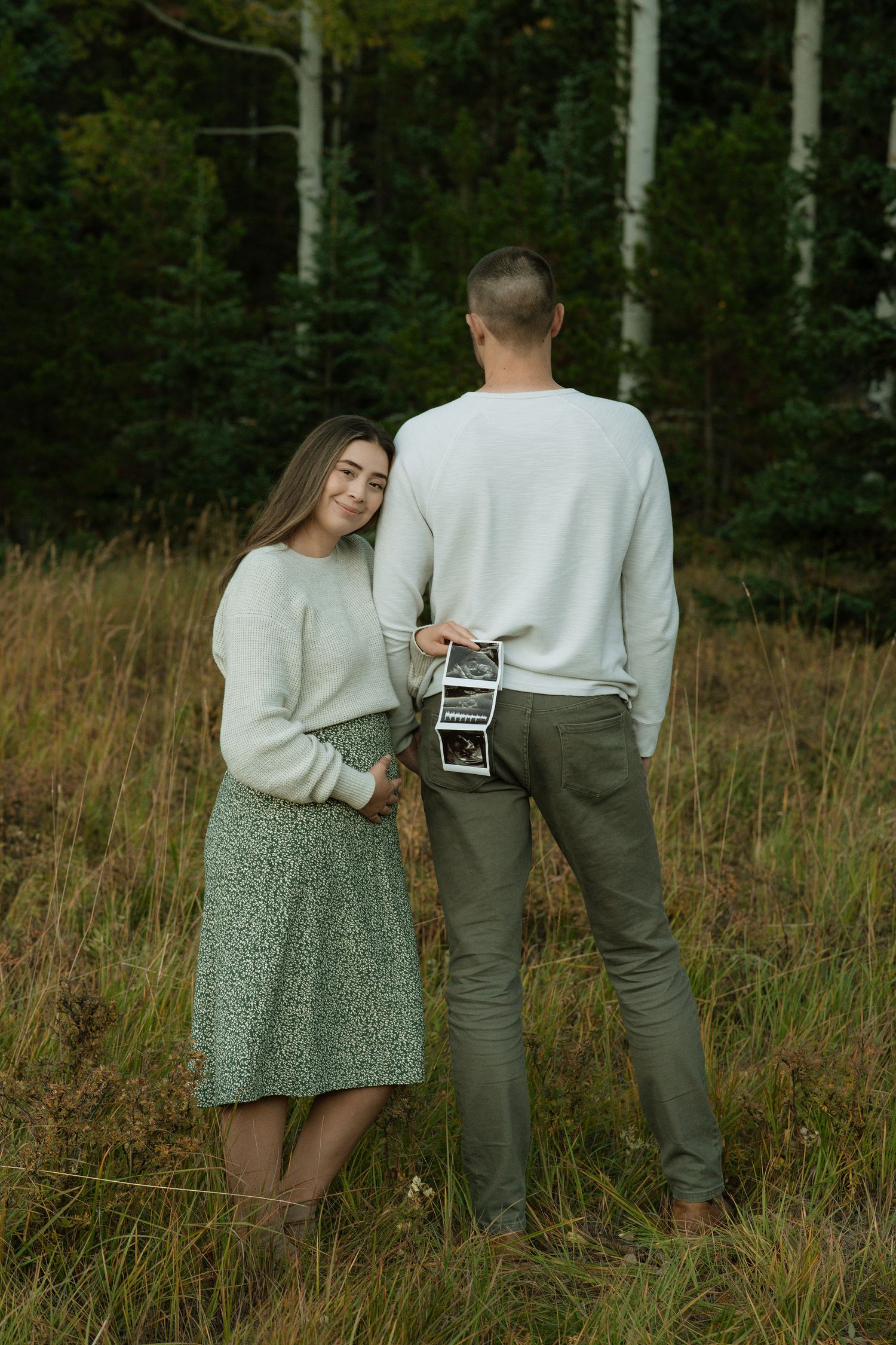 Couple holding ultrasound photos in a field, announcing pregnancy surrounded by autumn nature.