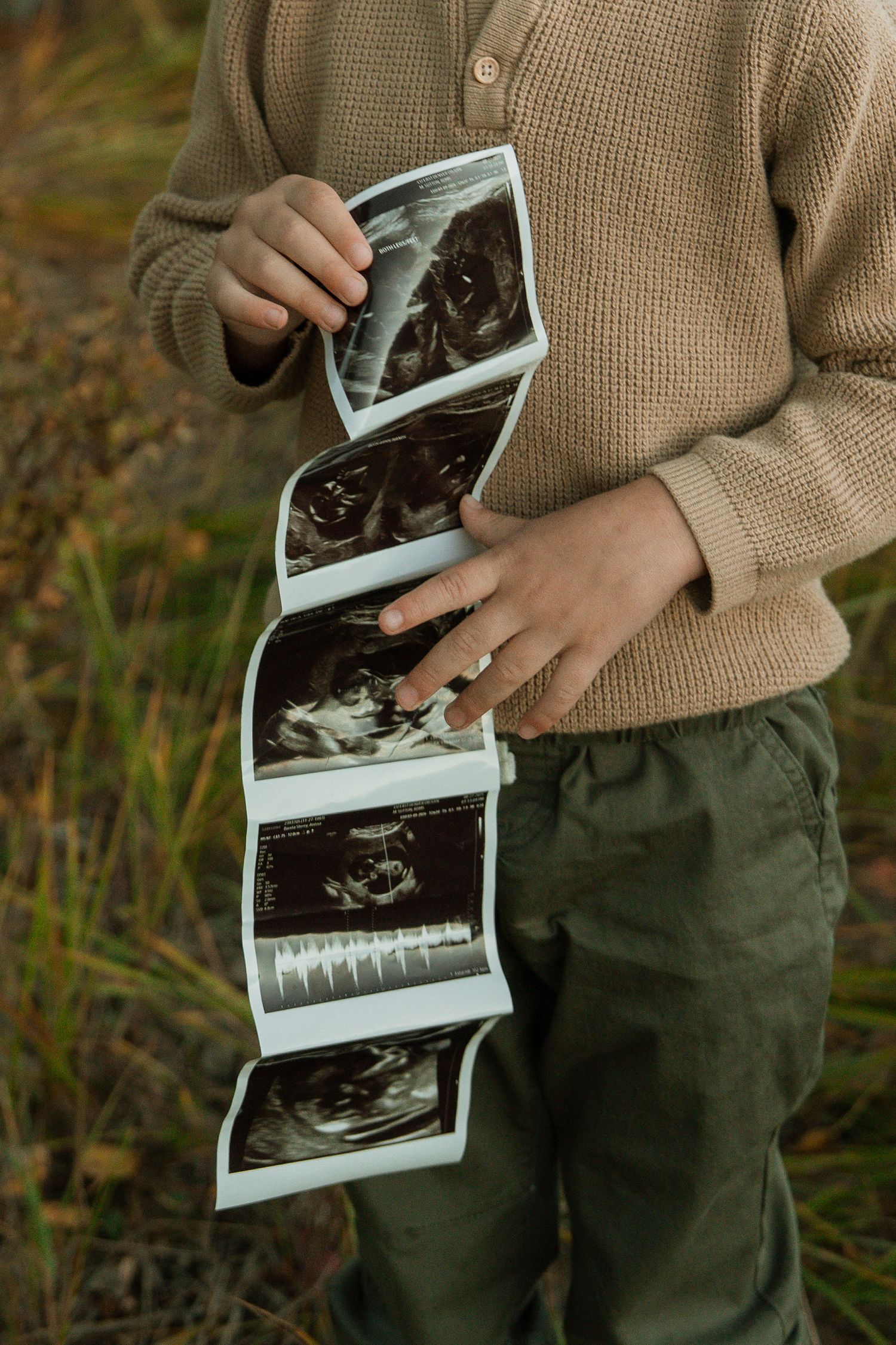 Pregnant woman in beige sweater holding ultrasound sonogram photos outdoors in nature, pregnancy announcement concept.