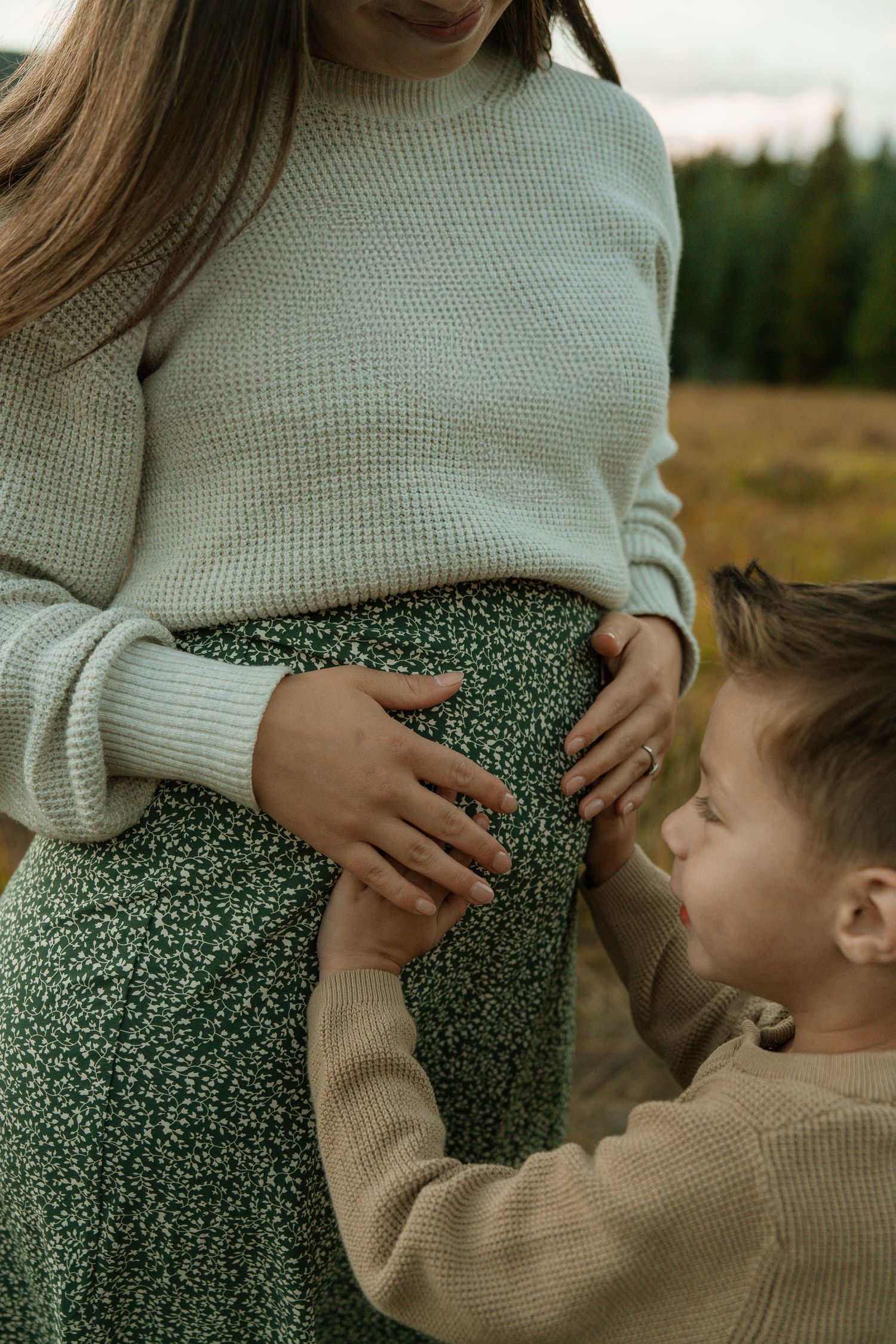 A young boy gently touches his pregnant mother's baby bump outdoors in a field, both wearing cozy knit sweaters.