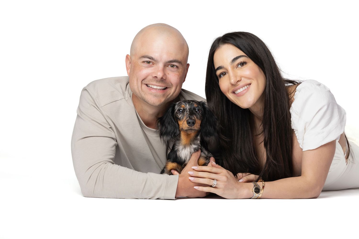 A smiling couple lying down together holding a small merle dachshund puppy on a white background.