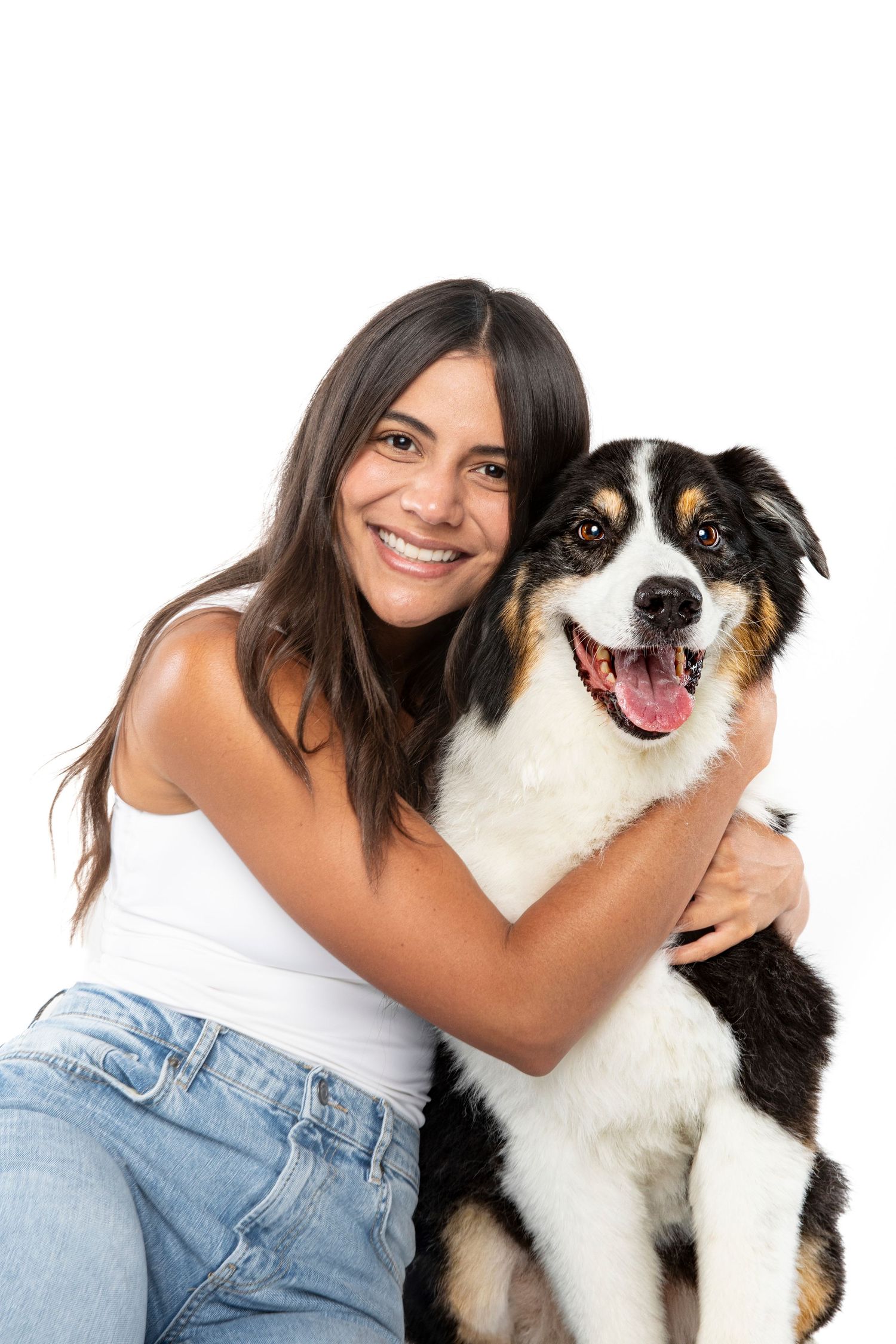 Smiling woman in white tank top hugging a black tri Australian Shepherd dog against a white background.