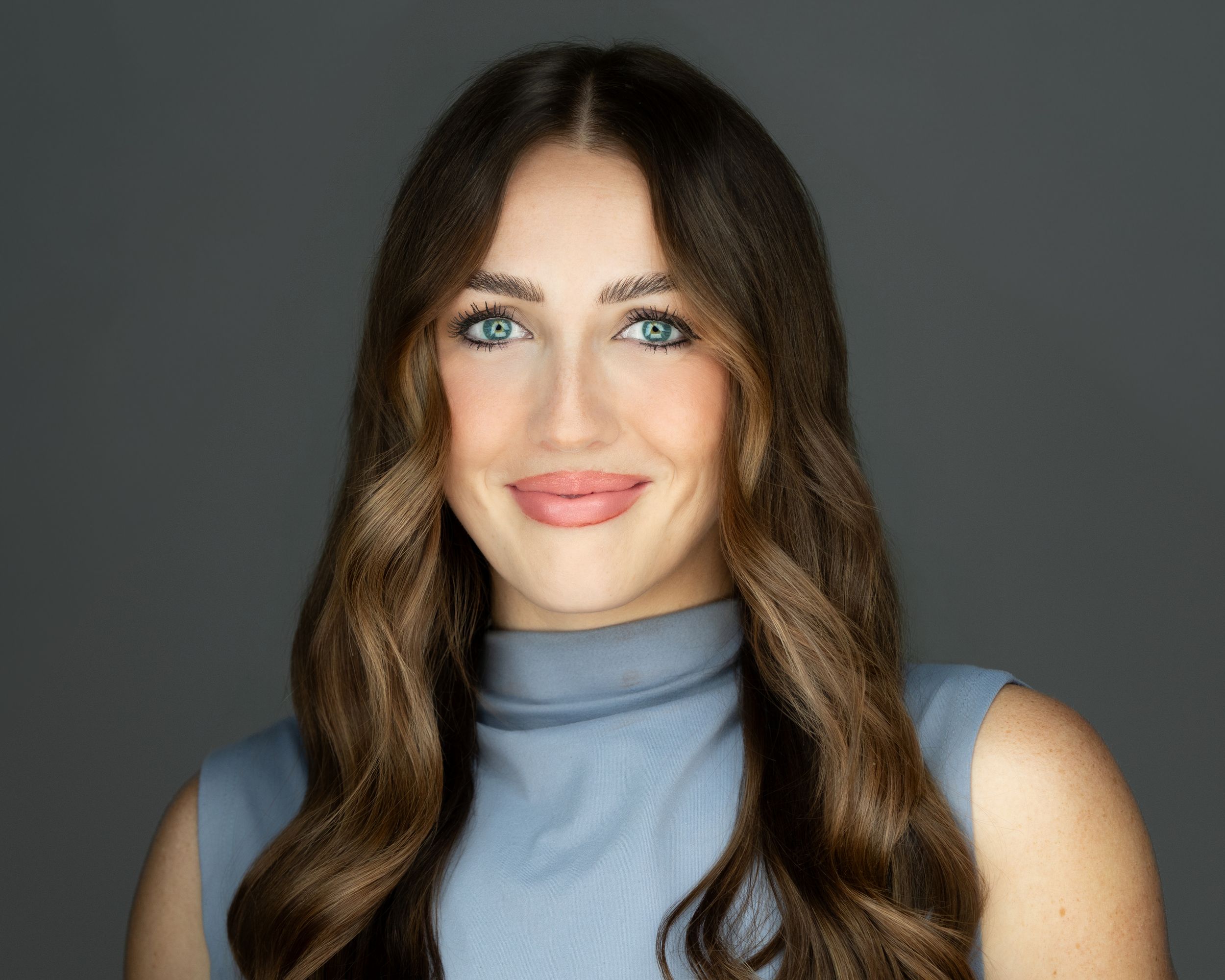 Professional headshot of brunette woman with blue eyes wearing light blue turtleneck top.