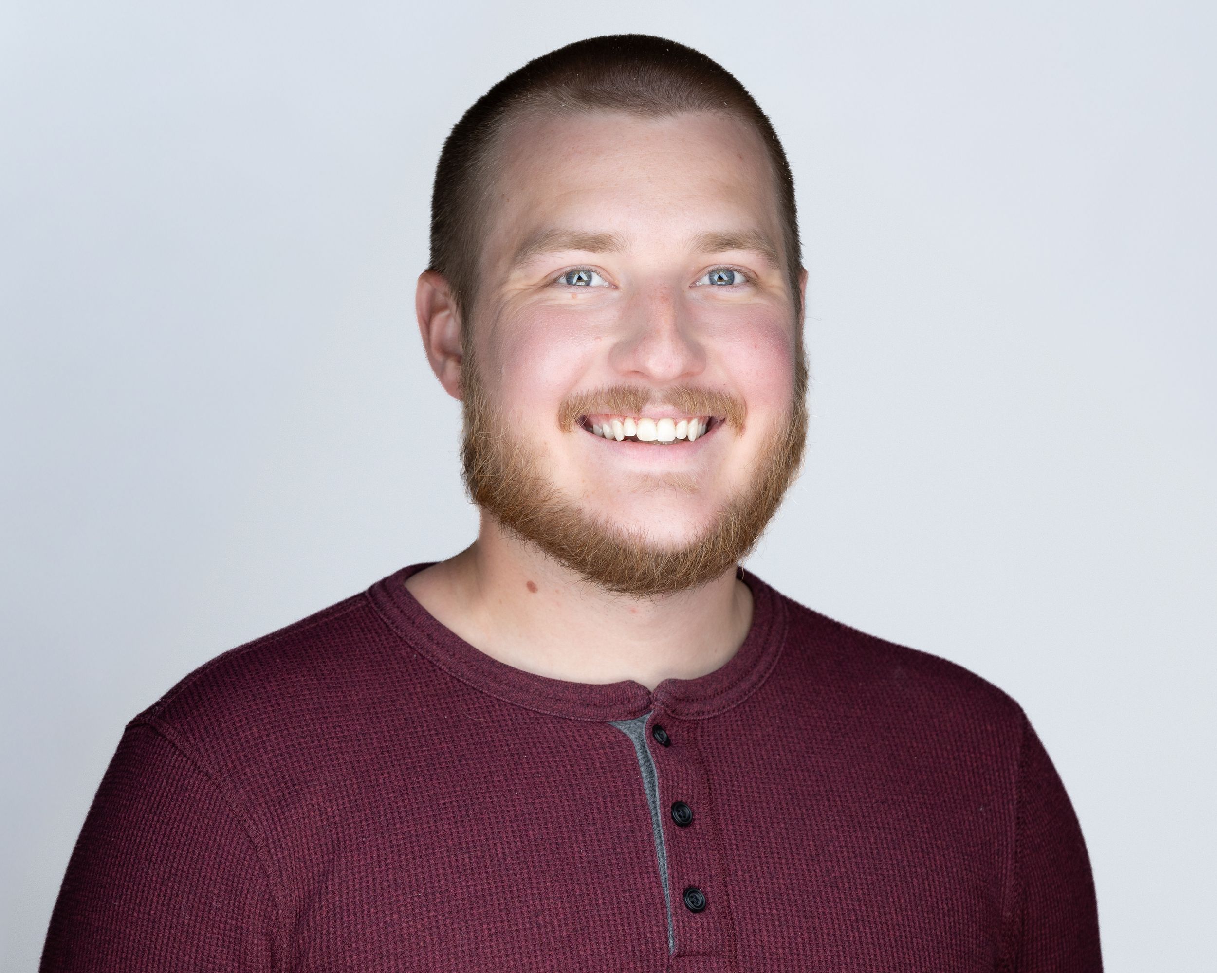 Professional headshot of smiling young man with beard wearing dark red henley shirt.