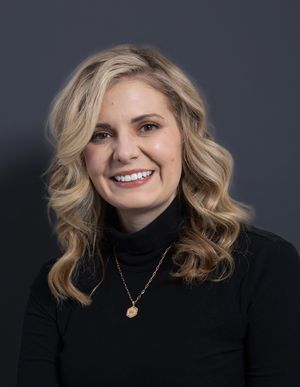 Professional headshot of a blonde woman in a black turtleneck with a gold necklace smiling against a dark background.