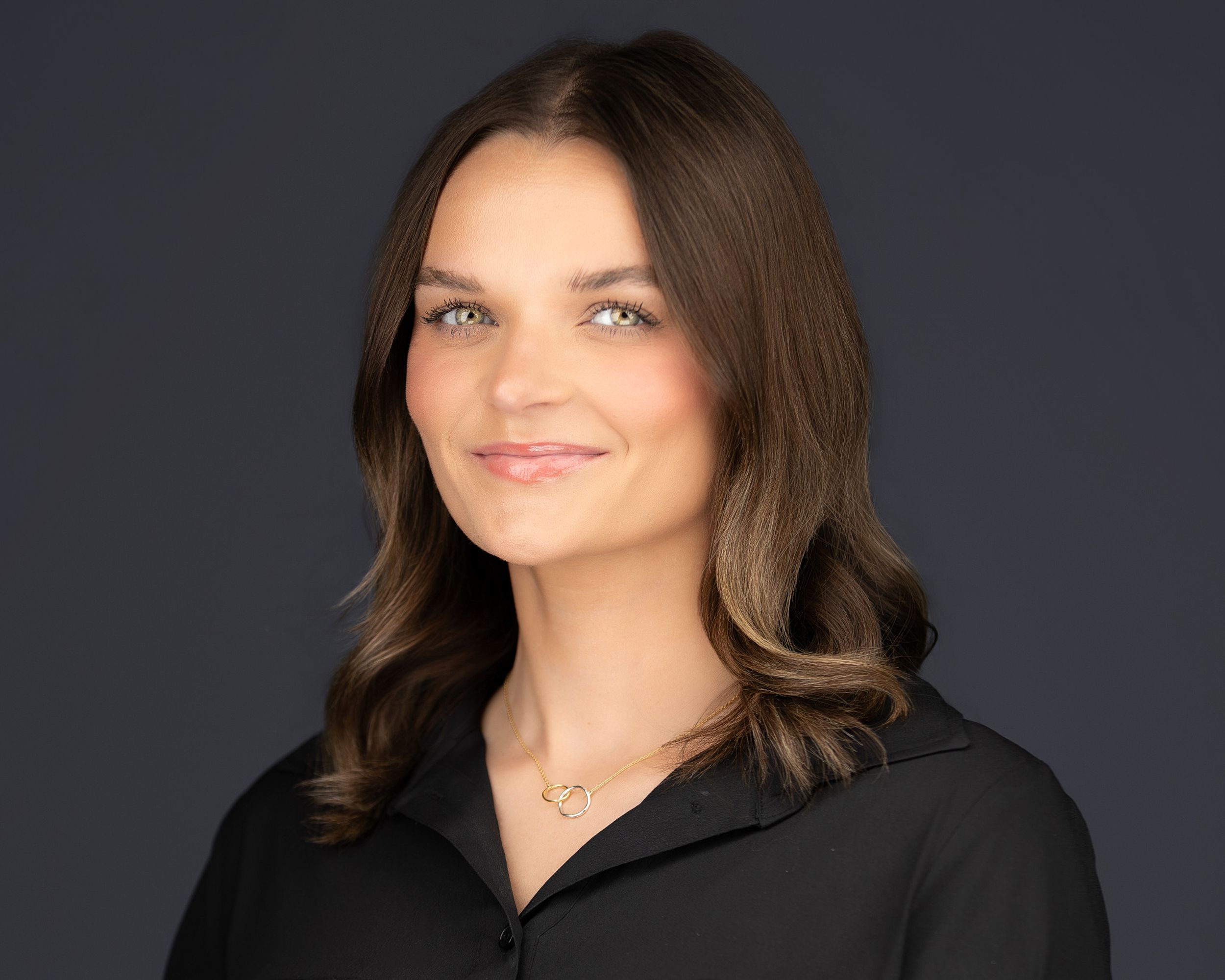 Professional headshot of a brunette woman in a black blouse against a dark gray studio background.