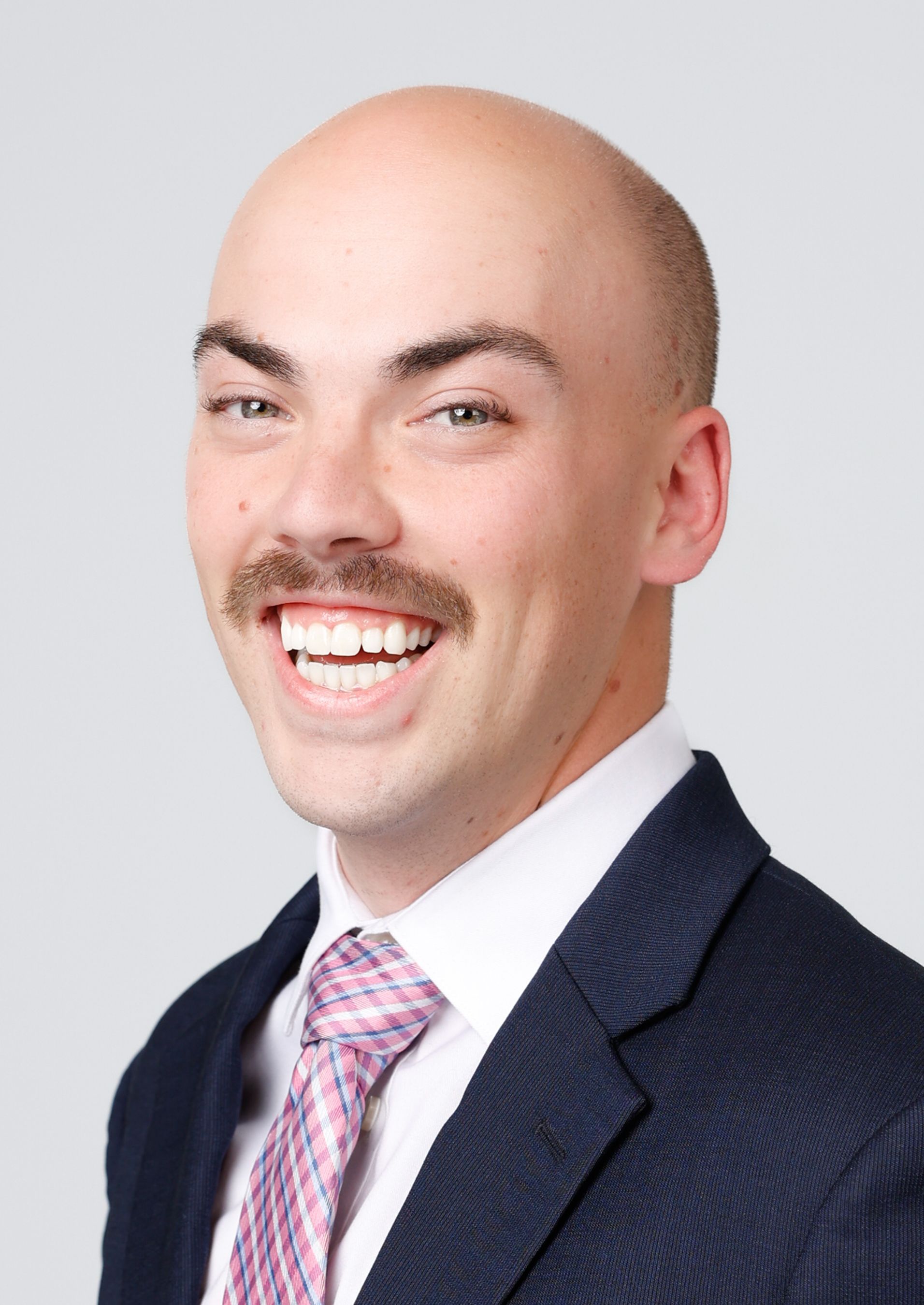 Professional headshot of a smiling bald man with a mustache wearing a navy suit and pink plaid tie.