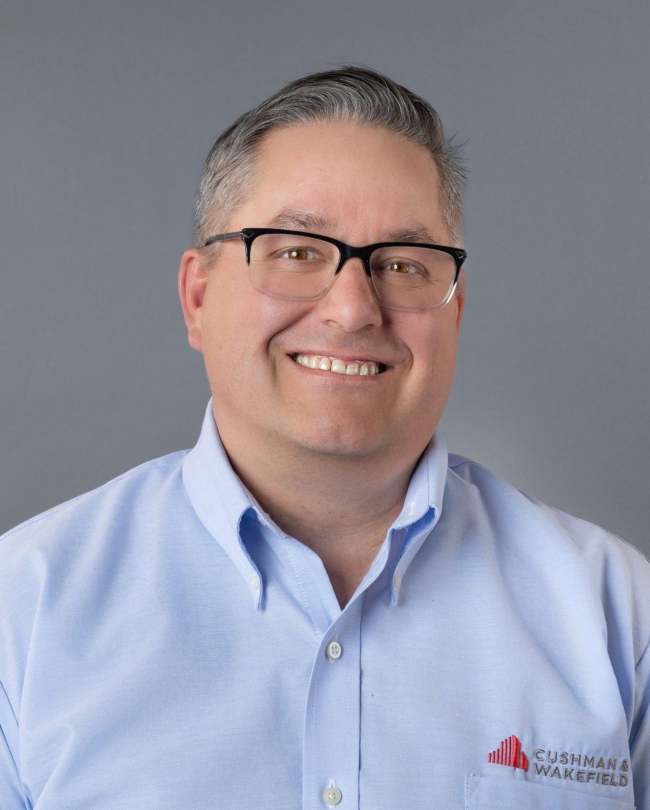Professional headshot of smiling man wearing glasses and light blue Cushman Wakefield shirt.