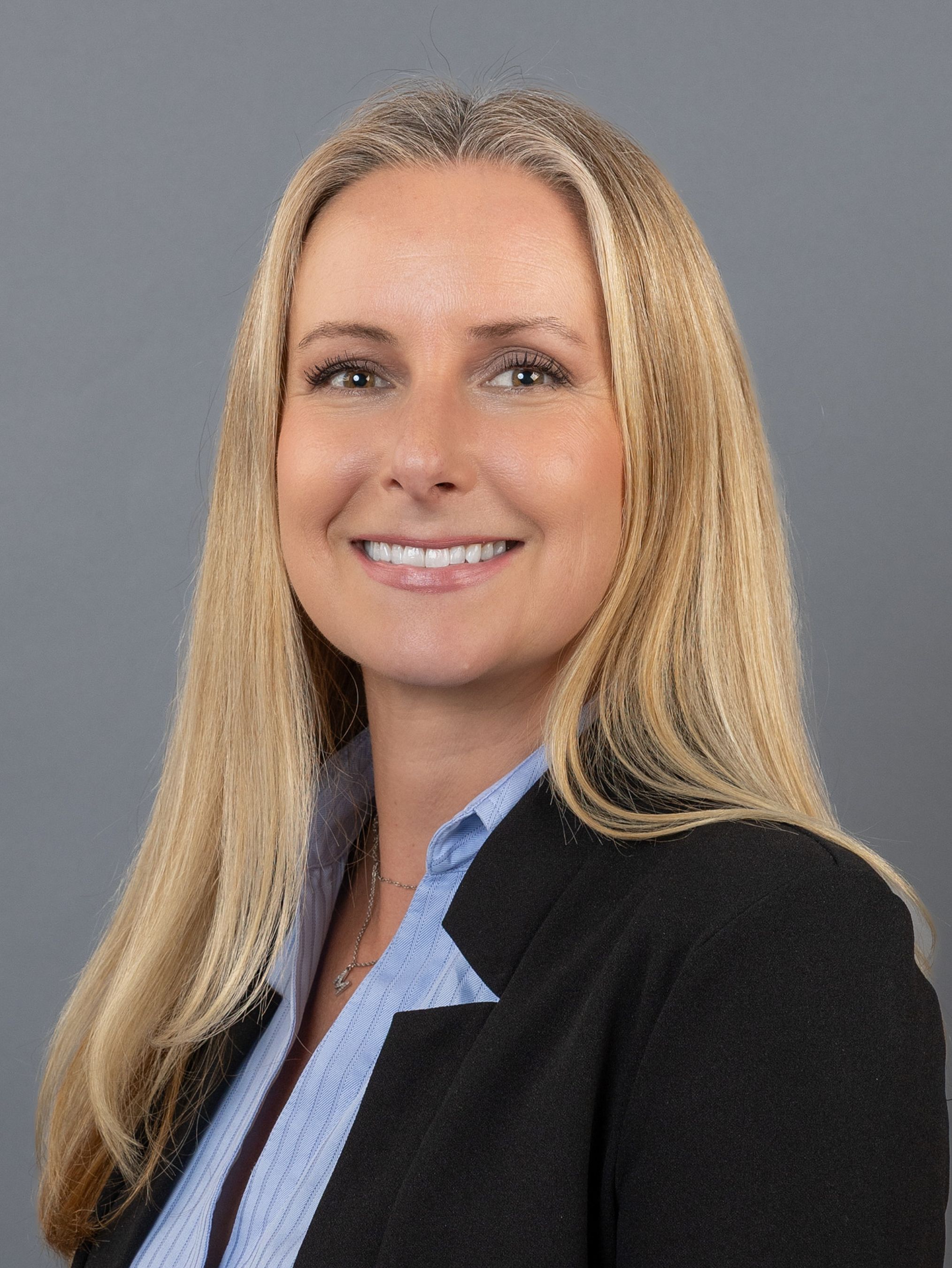 Professional headshot of a blonde woman in a black blazer and blue shirt smiling against a gray background.