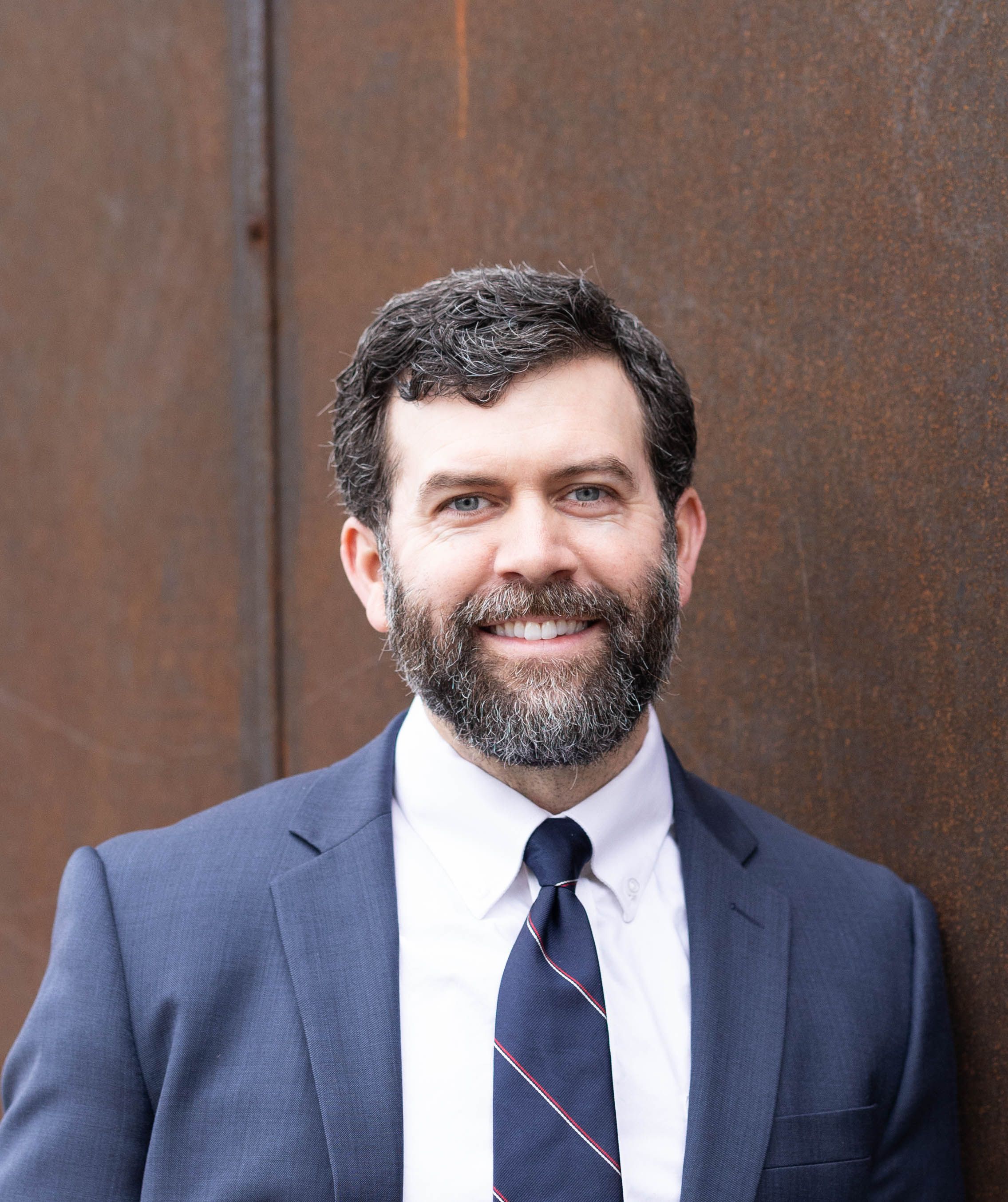 Professional headshot of a bearded man in a navy suit smiling against a rustic metal wall background.