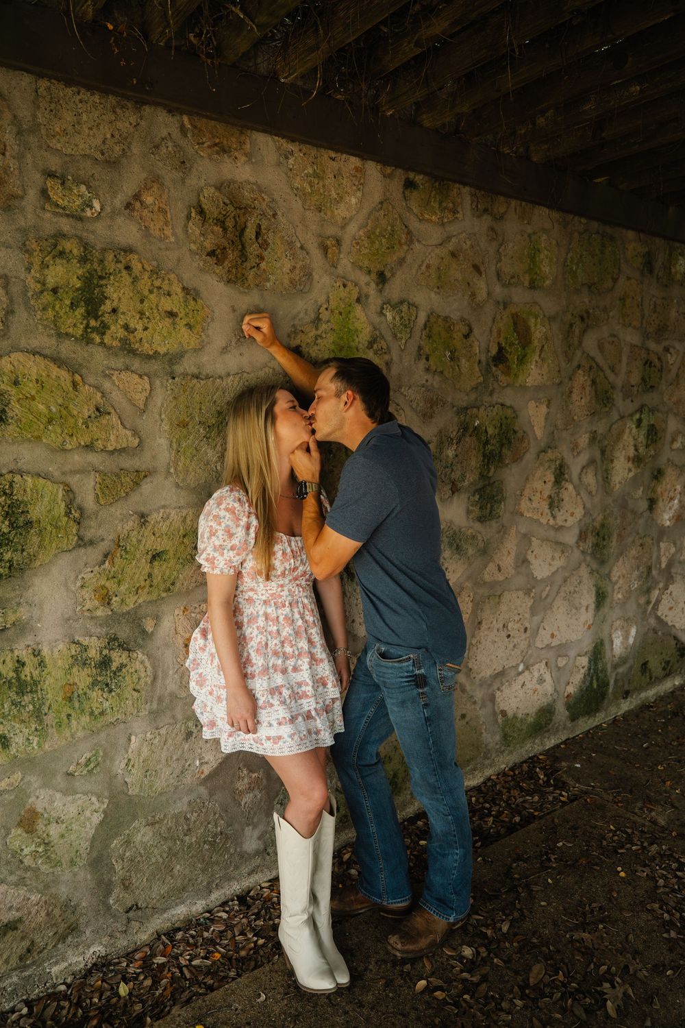 Couple shares a romantic kiss against a rustic moss-covered stone wall, woman wearing white cowboy boots.
