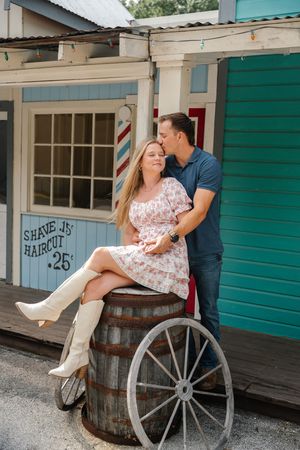 Woman in floral dress and white cowboy boots sits on a barrel as man kisses her cheek outside vintage shop.