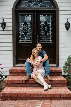 Happy engaged couple sits together on brick front porch steps of a charming home with elegant double doors.