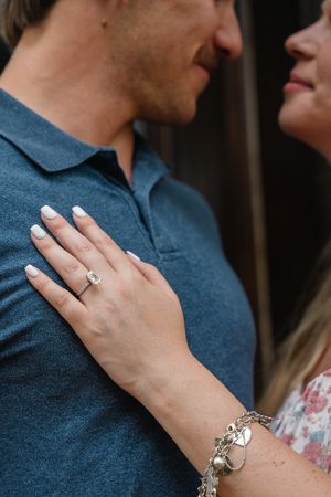 Close-up of woman's hand showing emerald-cut engagement ring and charm bracelet resting on man's blue shirt.