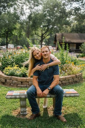 Smiling couple poses on a mosaic garden bench surrounded by colorful blooming flowers in a lush park.