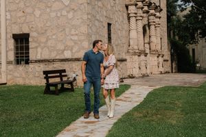 Engaged couple walks hand-in-hand along a stone path beside a historic mission-style building.