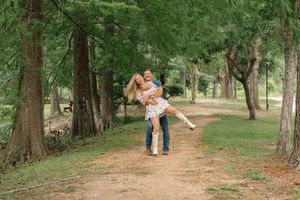 Joyful man lifts and spins his partner on a scenic dirt path surrounded by tall green trees.