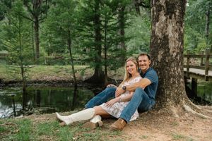 Happy couple sitting together against a tree by a pond in a green forest during engagement photos.