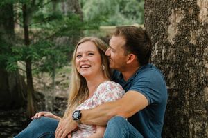 Couple laughing and cuddling against a tree in a lush forest during an outdoor engagement photo session.