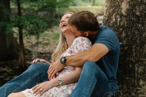 Joyful couple embracing and laughing together leaning against a tree in a wooded outdoor setting.
