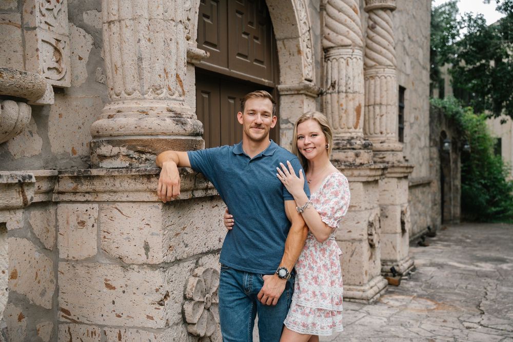 Smiling engaged couple posing together in front of ornate stone architecture during engagement session.