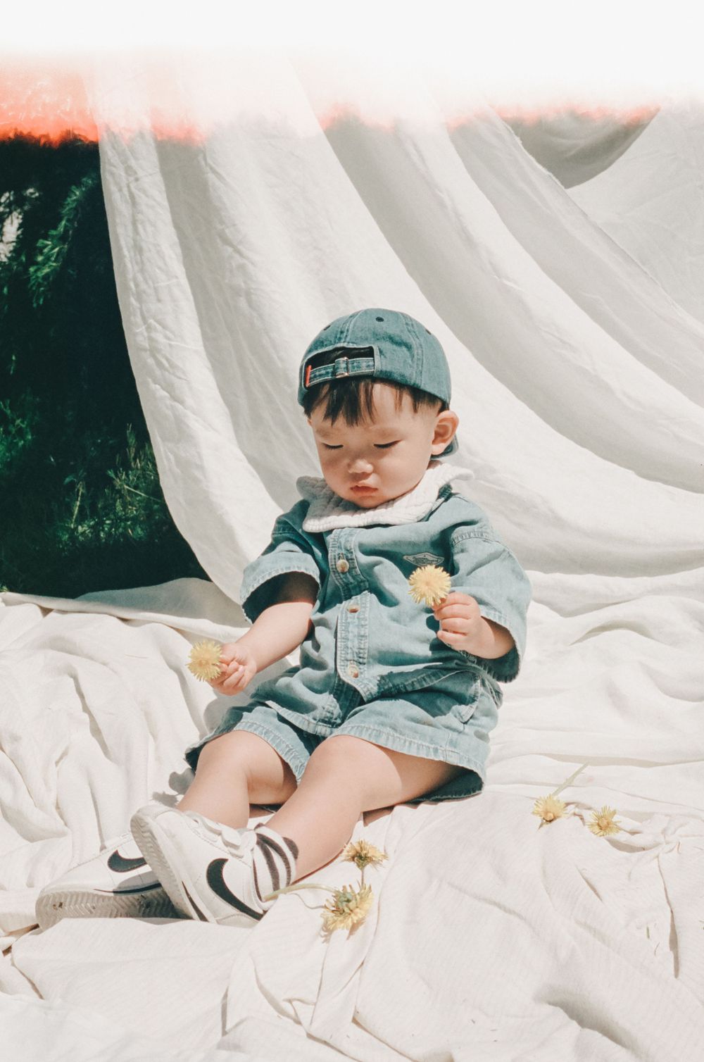 A cute baby boy in a denim romper and baseball cap sits on white fabric, holding small yellow flowers outdoors.
