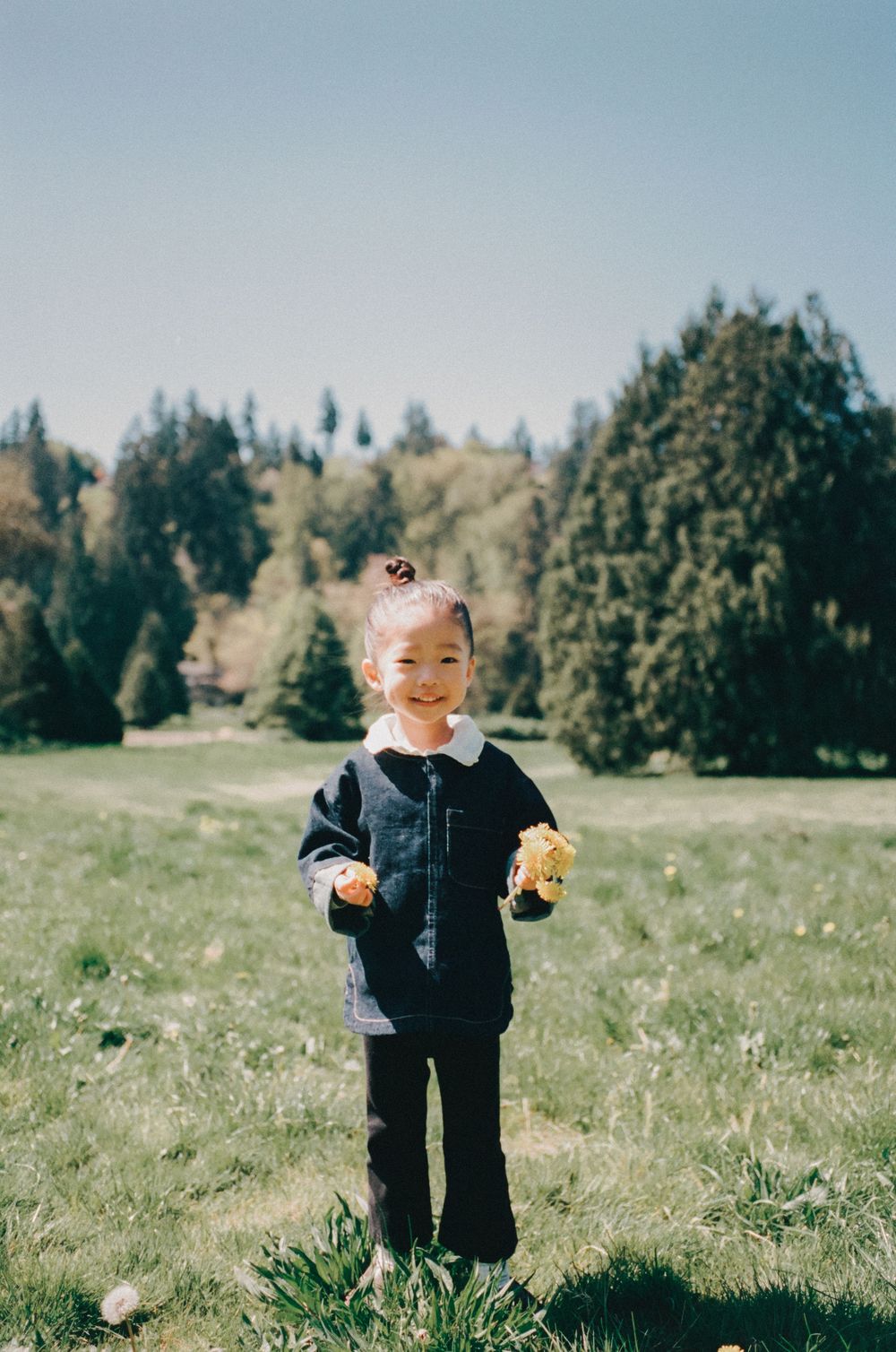 Young girl in black jacket holding yellow dandelions smiling in a green meadow with trees in background.