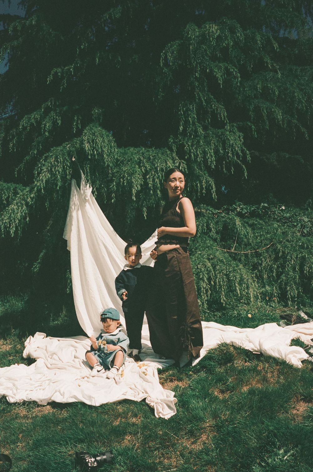 A woman and toddler pose outdoors on white fabric near a draped cloth backdrop with lush green weeping trees behind them.