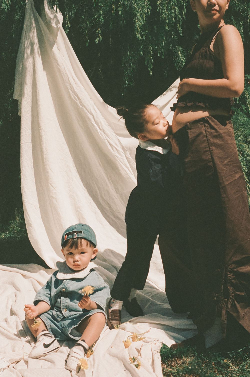 Two young children and a woman pose outdoors near a white fabric backdrop with willow trees in background.