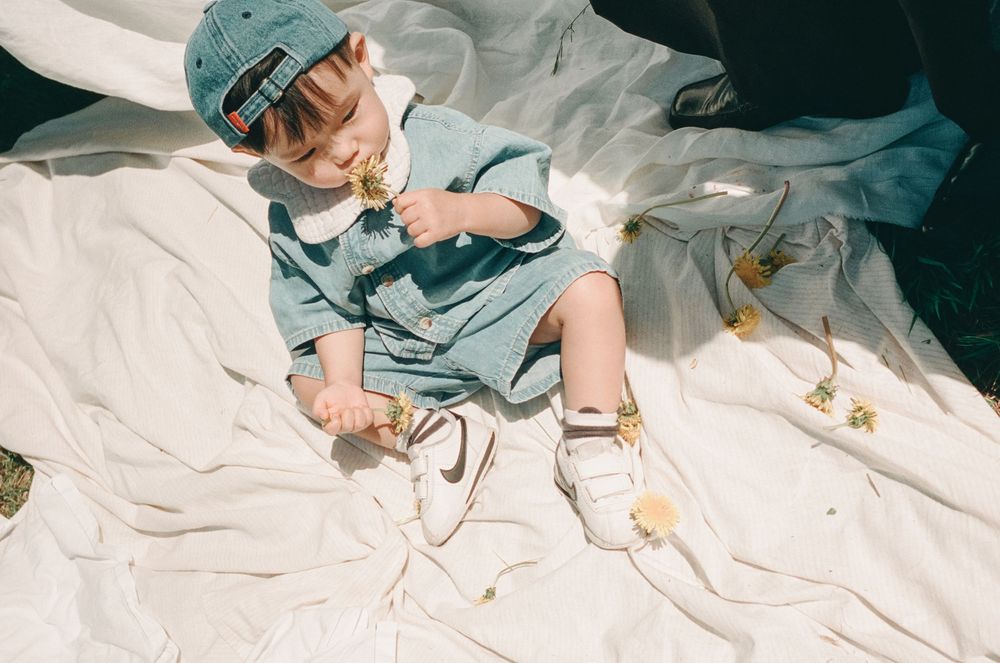 A toddler in a blue denim outfit and cap sits on white fabric, playing with white sneakers and daisy accessories.