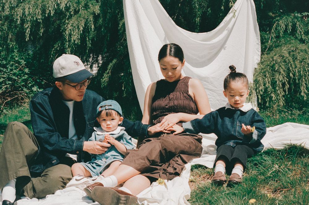 Family of four enjoying a picnic outdoors, sitting on a white blanket surrounded by lush green trees.